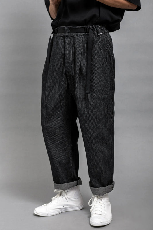 Men's dark grey pleated trousers with a relaxed fit, worn cuffed at the ankle, paired with casual white high-top sneakers and styled with a black belt.