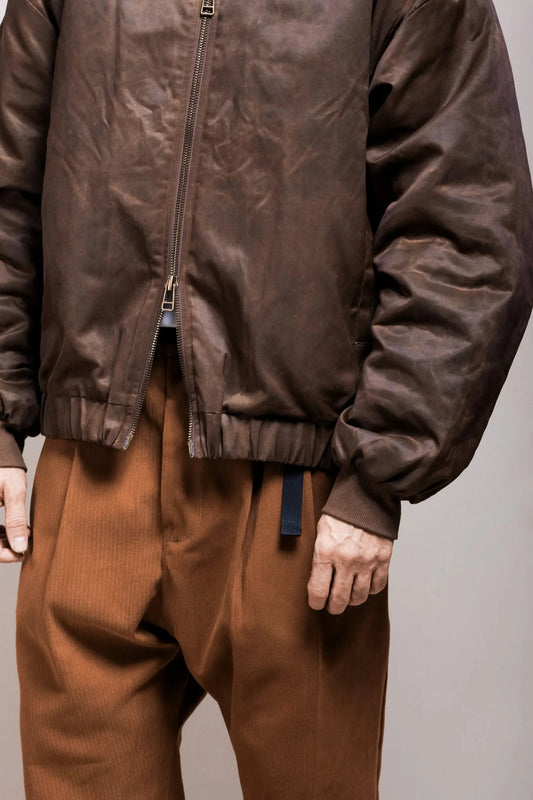 Close-up of a person wearing a chocolate brown bomber-style jacket with a zip closure and elastic cuffs, paired with loose-fitting brown pleated pants in a relaxed menswear style.