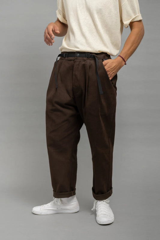 Man wearing relaxed fit pleated chocolate brown pants with a black belt, paired with a cream short-sleeve shirt and white sneakers against a plain gray background.