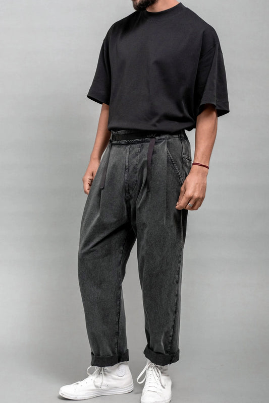 Man wearing relaxed fit pleated black wash double-knit pants paired with a loose black t-shirt and white sneakers against a plain gray background