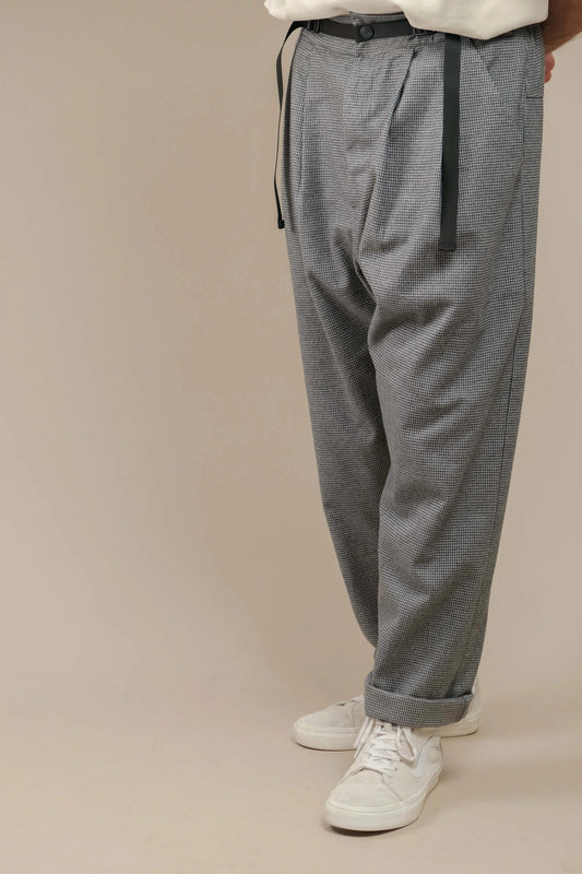 Man wearing relaxed fit grey houndstooth pants with pleats and cuffed hems paired with white sneakers against a neutral background.