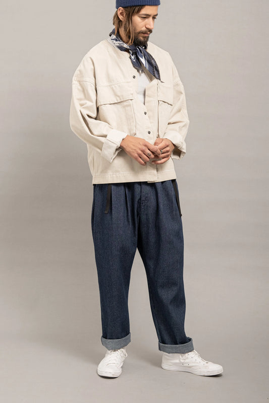 Man wearing relaxed fit indigo pleated pants paired with a loose off-white jacket, white sneakers, and a navy patterned bandana around the neck.