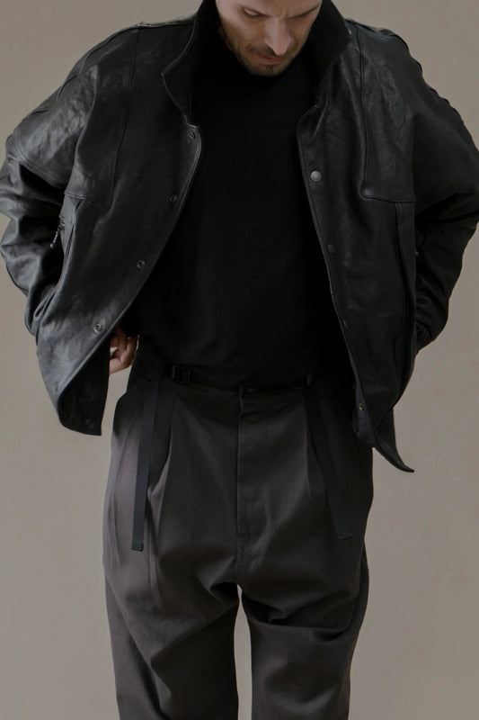 Man wearing a relaxed-fit black leather jacket over a black shirt paired with dark high-waisted pleated pants with belt straps, showcasing a minimalist, utility-inspired look.