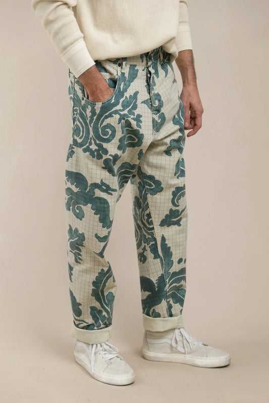 Man wearing relaxed fit pants with a blue-green floral and scroll pattern on a light background, paired with white sneakers and a cream-colored sweater.