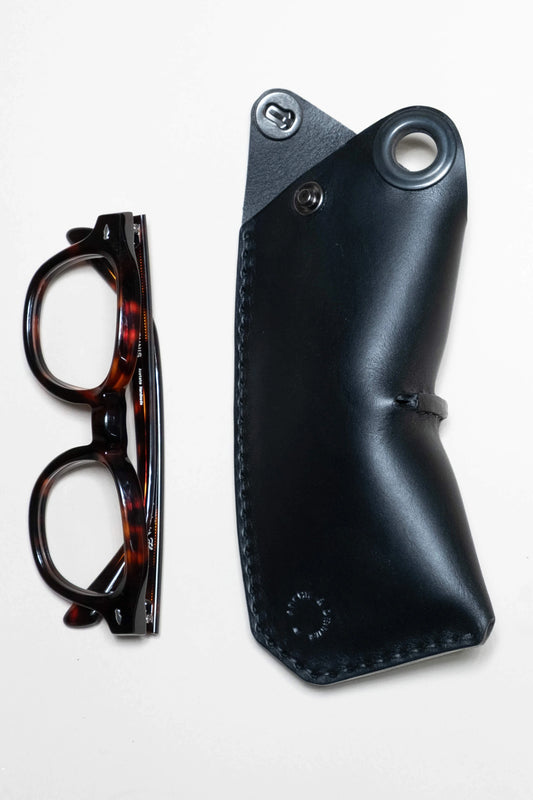 Tortoiseshell glasses next to a sleek black leather glasses case with stitched edges and metal snap closure, designed for durability and style.