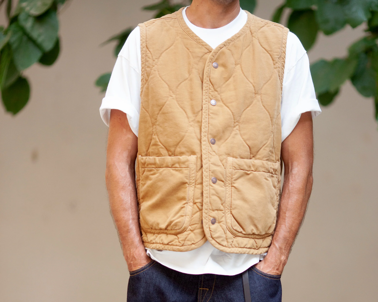 Man wearing a quilted tan vest with snap buttons over a white short-sleeve t-shirt, hands in pockets, standing against a blurred natural background.