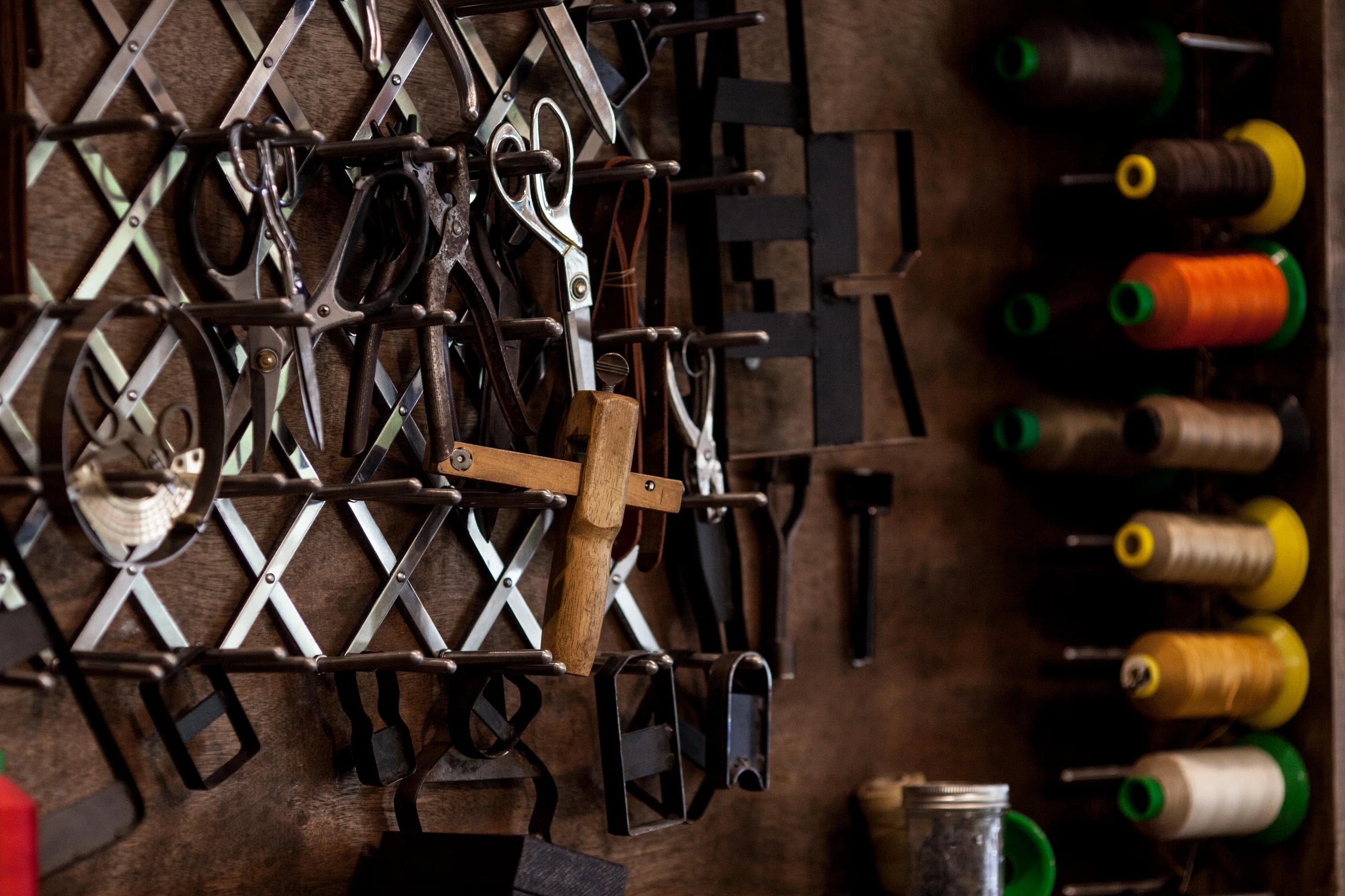 Wall-mounted metal rack holding various sewing tools including scissors, thread spool holder with multiple colored threads, measuring tape, and a wooden tool in a dimly lit workshop setting.