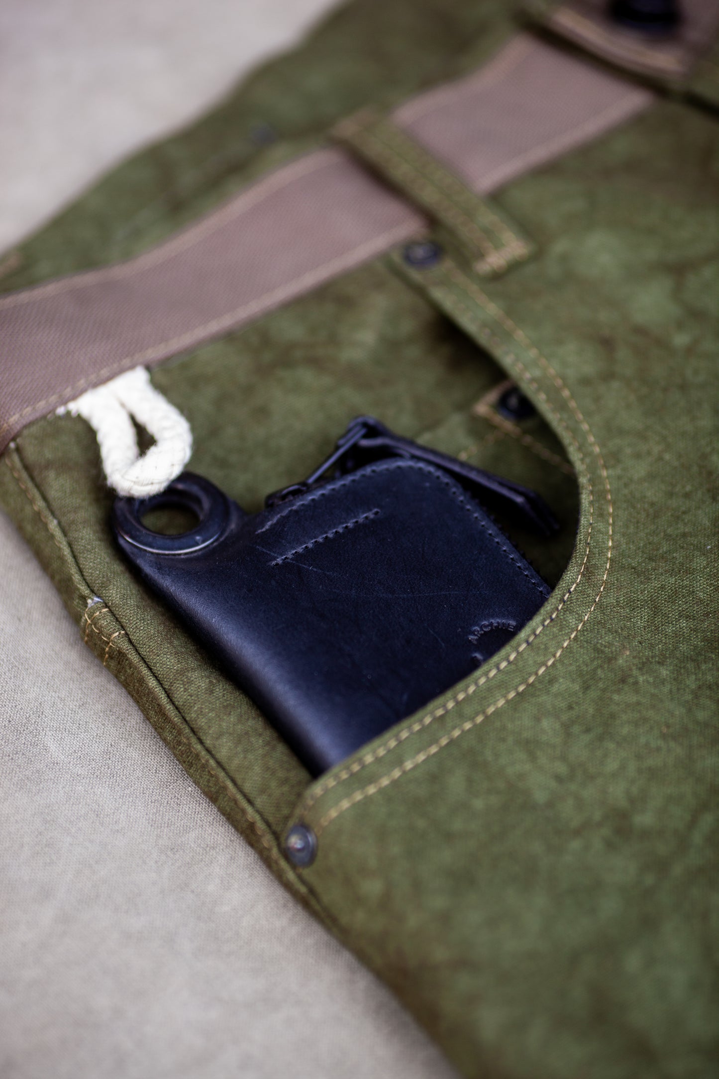 Black leather zipper wallet with a loop attached, partially tucked into the front pocket of olive green utility-style pants with detailed stitching.