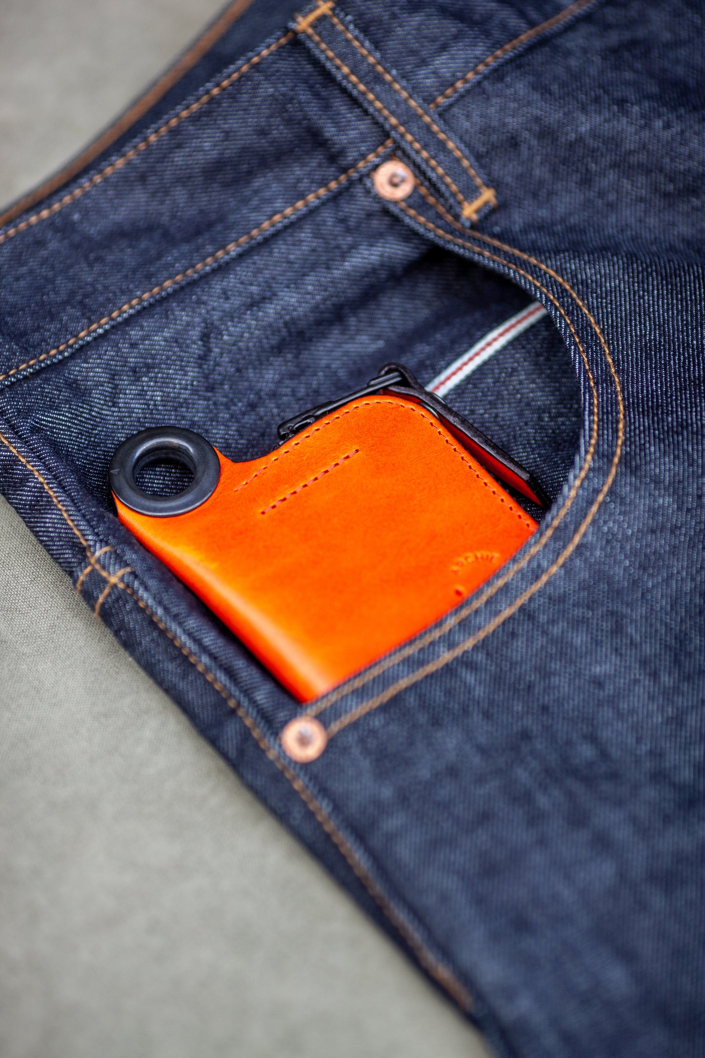 Orange leather zipper wallet with a circular keyring detail, tucked into the front pocket of dark blue denim jeans with visible orange stitching.
