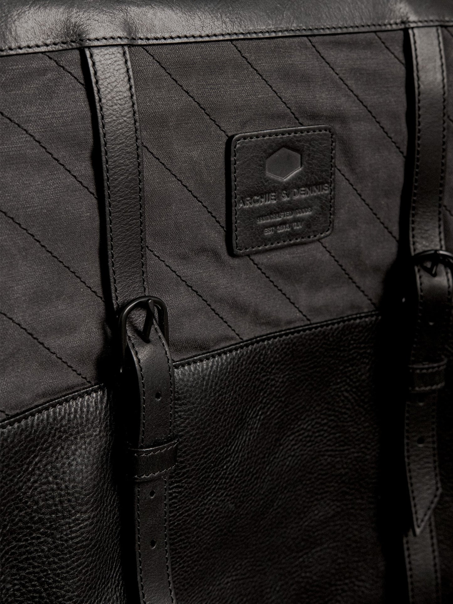 Close-up of a black handmade bag featuring textured leather, durable stitched fabric, adjustable leather straps, and a branded label, highlighting craftsmanship and utility style.