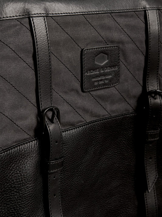 Close-up of a black handmade bag featuring textured leather, durable stitched fabric, adjustable leather straps, and a branded label, highlighting craftsmanship and utility style.