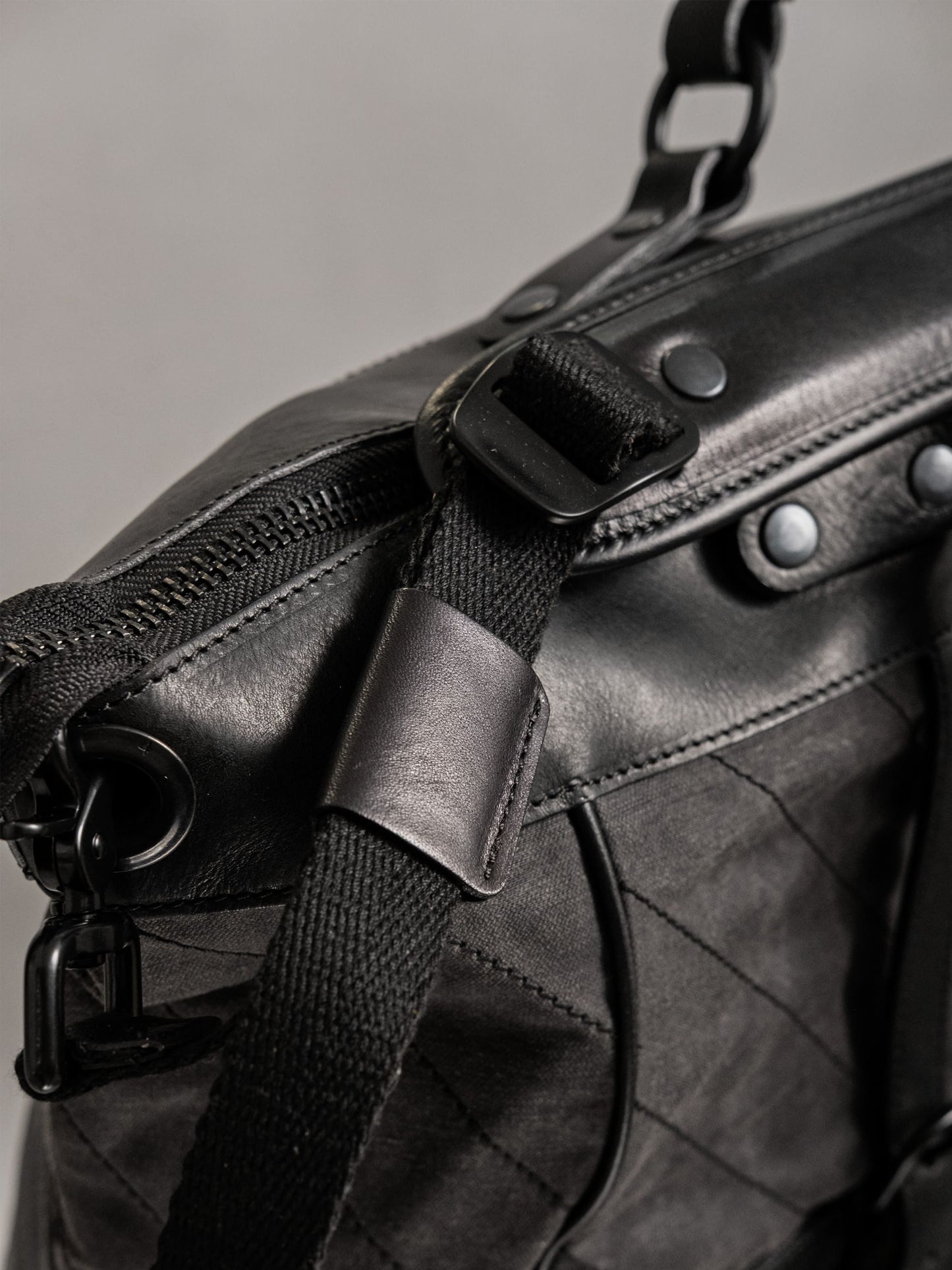 Close-up of a black handcrafted bag featuring textured leather, durable fabric strap with adjustable buckle, and metal hardware detailing for a sleek, utility-inspired look.