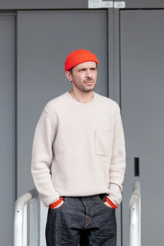 Man wearing a cream-colored ribbed sweater with a chest pocket, paired with dark denim jeans and a bright orange beanie, standing against a gray urban background.