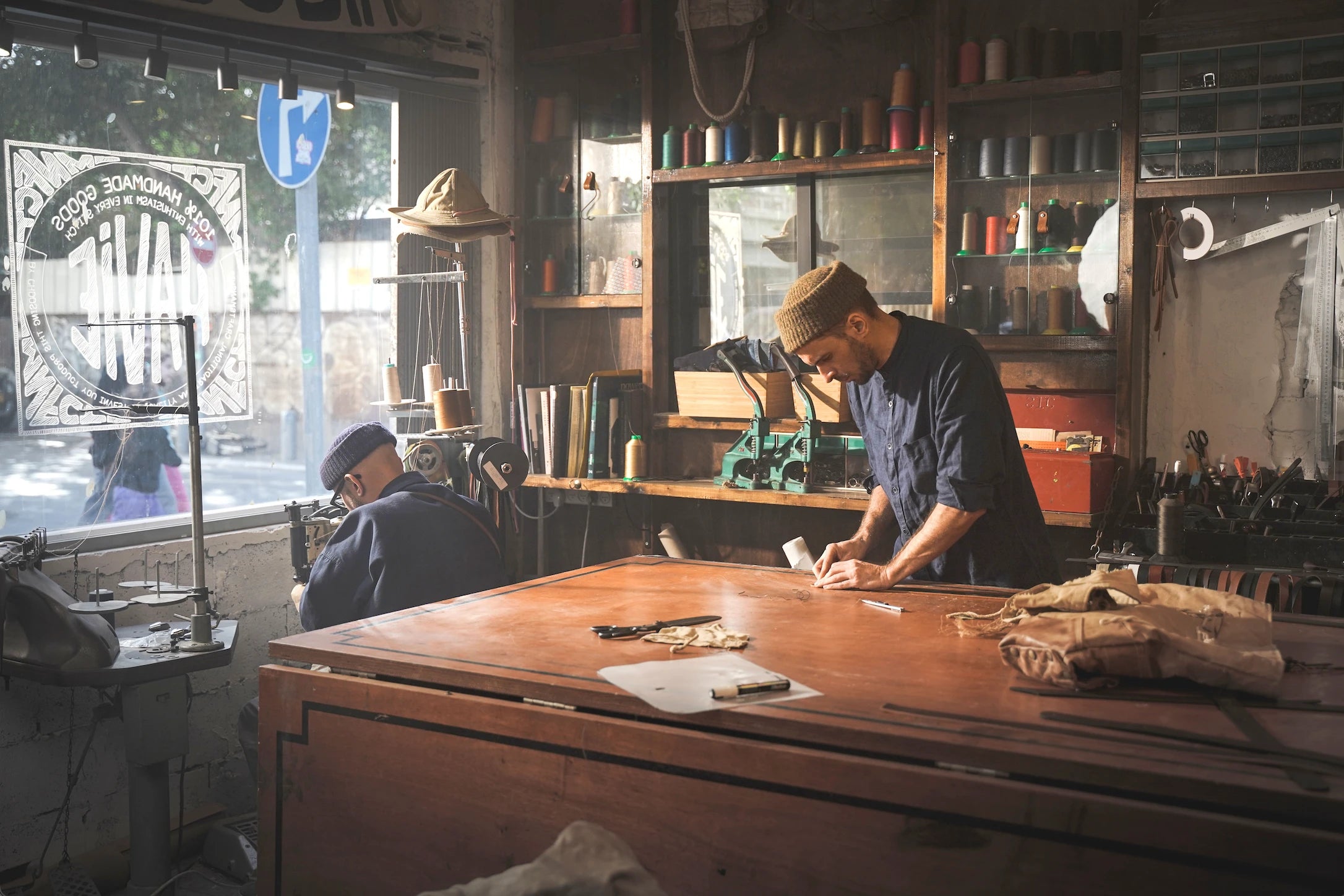 Two men working in a rustic tailor shop filled with sewing supplies, fabric, and vintage tools bathed in natural light from a large window.