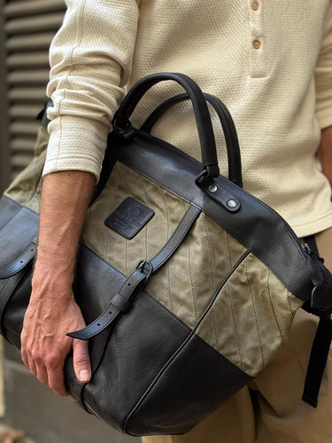 Man holding a stylish two-tone tote bag featuring black leather and olive quilted fabric with buckle detail, paired with a casual beige long sleeve top and pants.