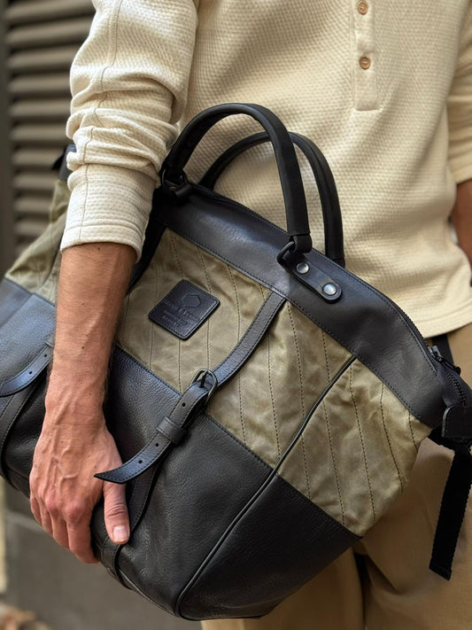Man holding a stylish two-tone tote bag featuring black leather and olive quilted fabric with buckle detail, paired with a casual beige long sleeve top and pants.