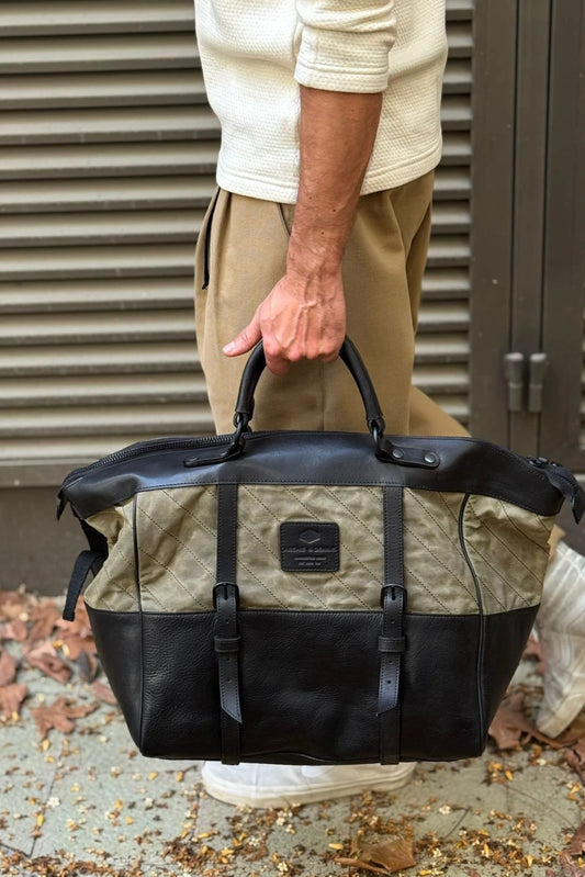 Man holding a sturdy handmade weekend bag with quilted olive green fabric and black leather accents, showcasing a sleek utility-inspired design.