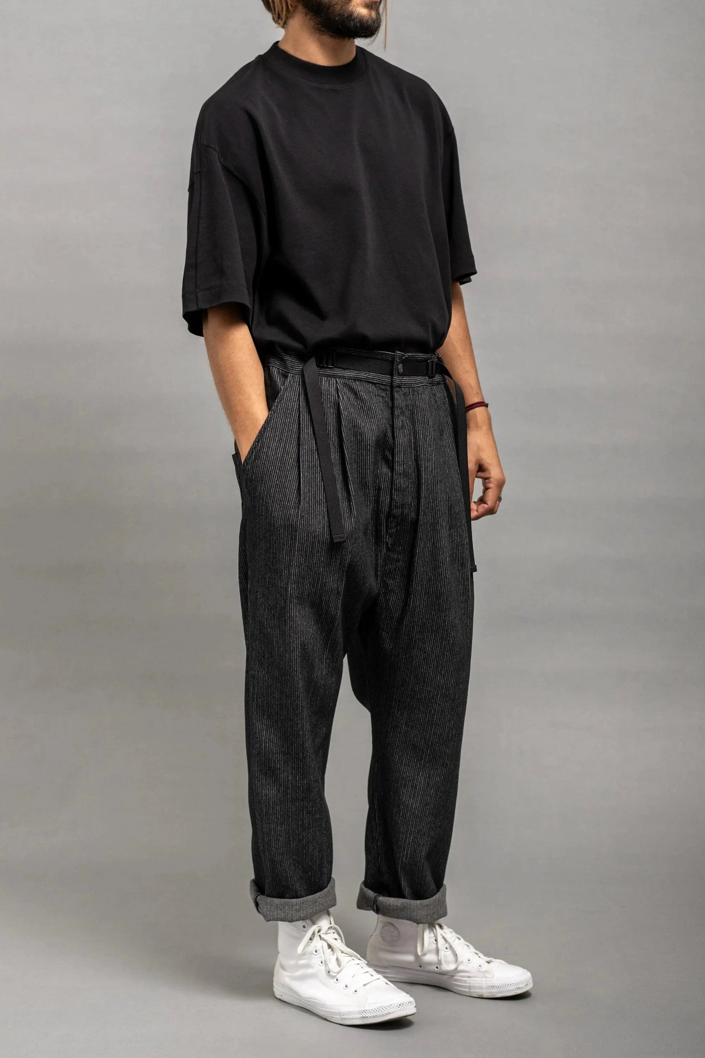 Man wearing relaxed fit pleated dark grey pants with a fabric belt, paired with a loose black t-shirt and white high-top sneakers against a plain gray background.