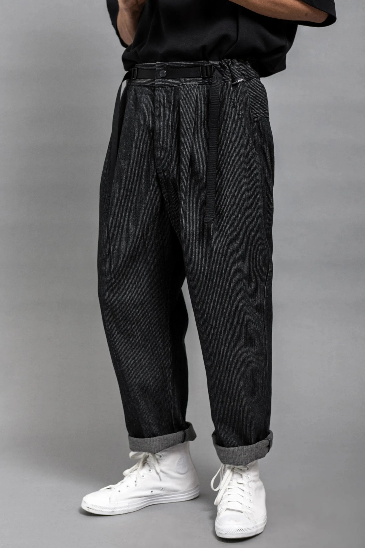 Men's dark grey pleated trousers with a relaxed fit, worn cuffed at the ankle, paired with casual white high-top sneakers and styled with a black belt.