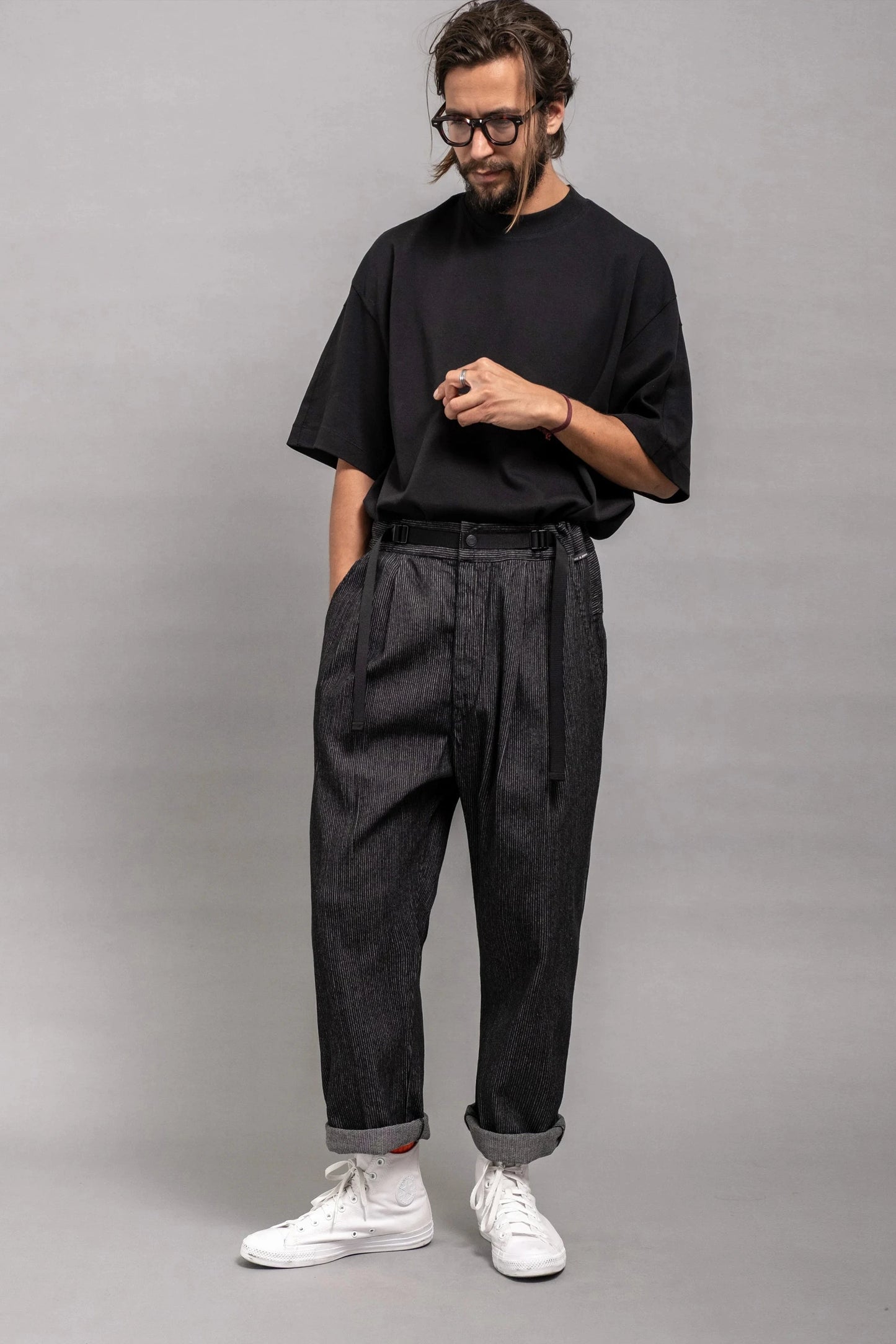 Man wearing relaxed fit pleated dark grey pants with rolled cuffs, paired with a loose black t-shirt and white sneakers, styled with glasses and a beard against a plain gray background.