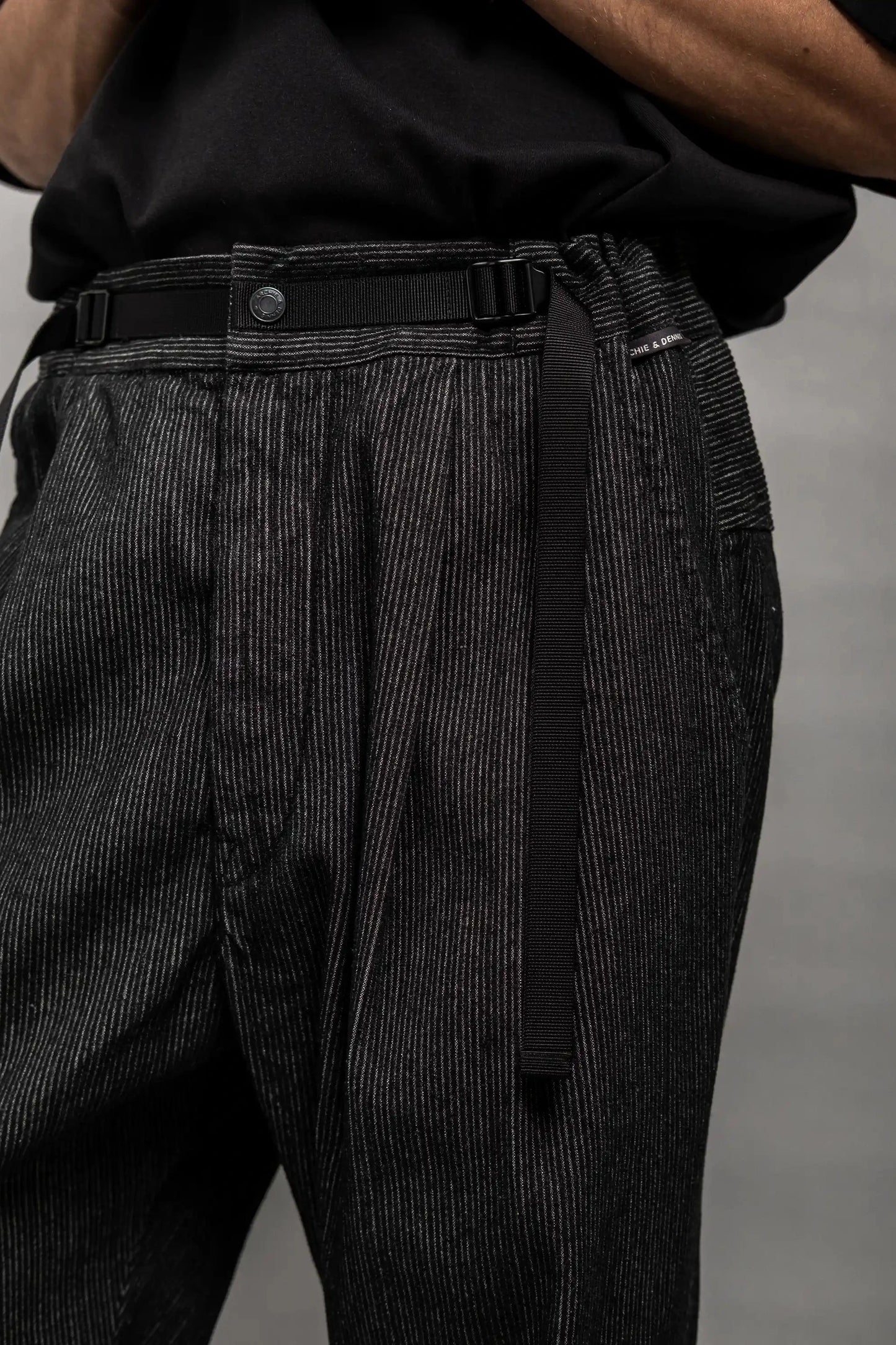 Close-up of black striped relaxed fit pants with a built-in adjustable belt, worn with a tucked-in black shirt, showcasing detailed craftsmanship and a subtle textured pattern.