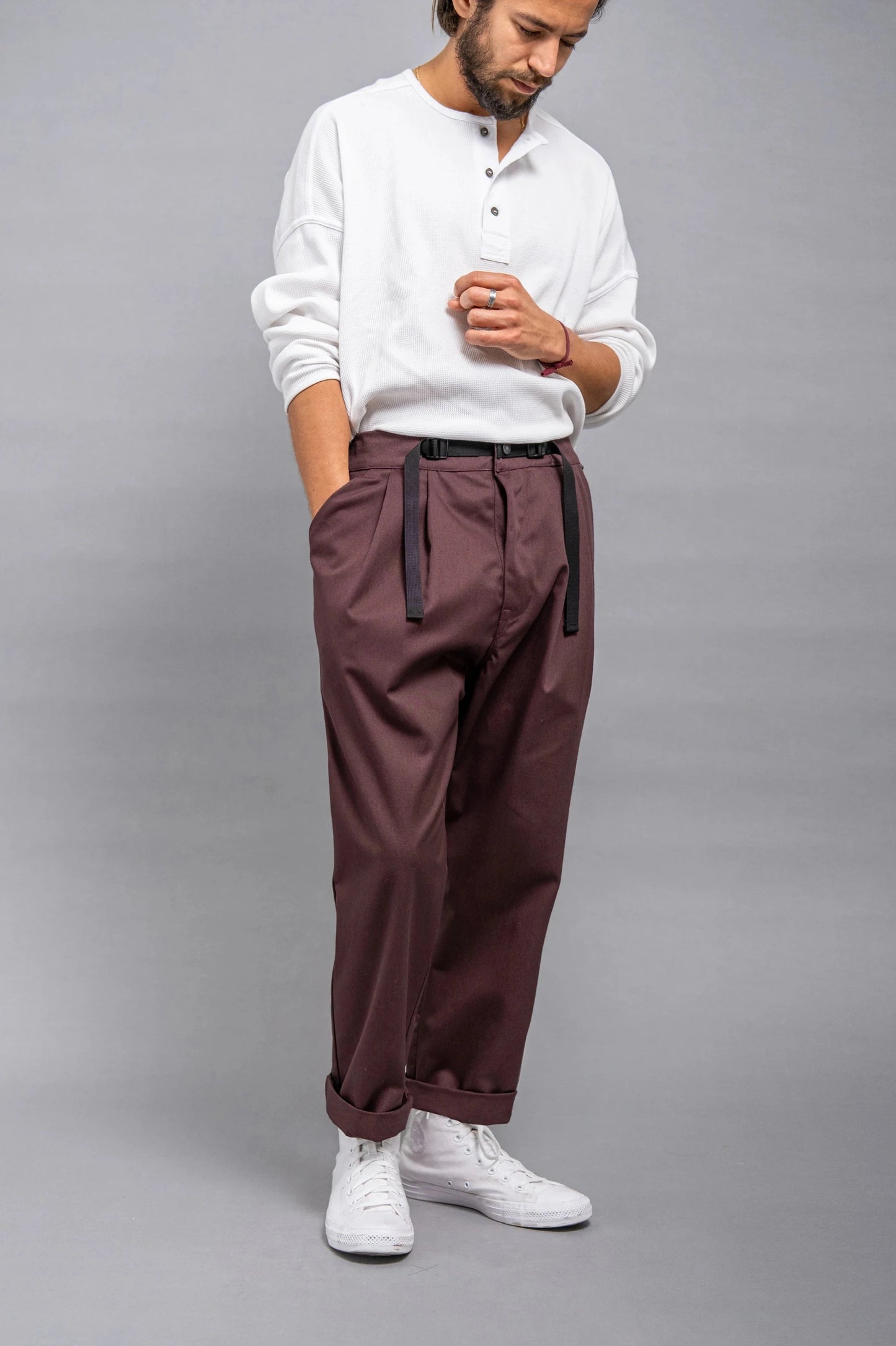 Man wearing relaxed fit pleated burgundy pants with black belt and rolled cuffs paired with a white long-sleeve henley shirt and white sneakers against a gray background.