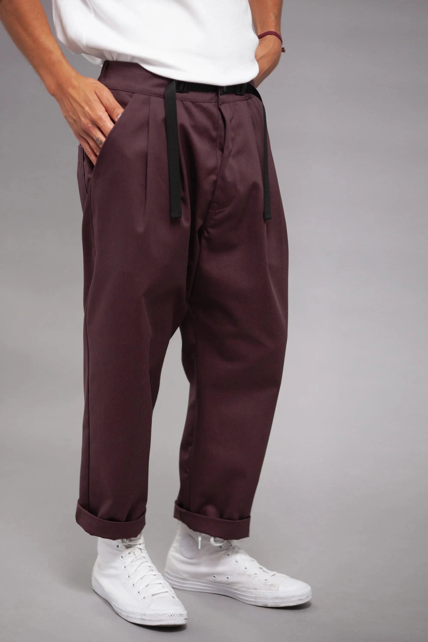 Man wearing relaxed fit pleated burgundy pants with a black drawstring and cuffed hems, paired with white high-top sneakers against a neutral background.