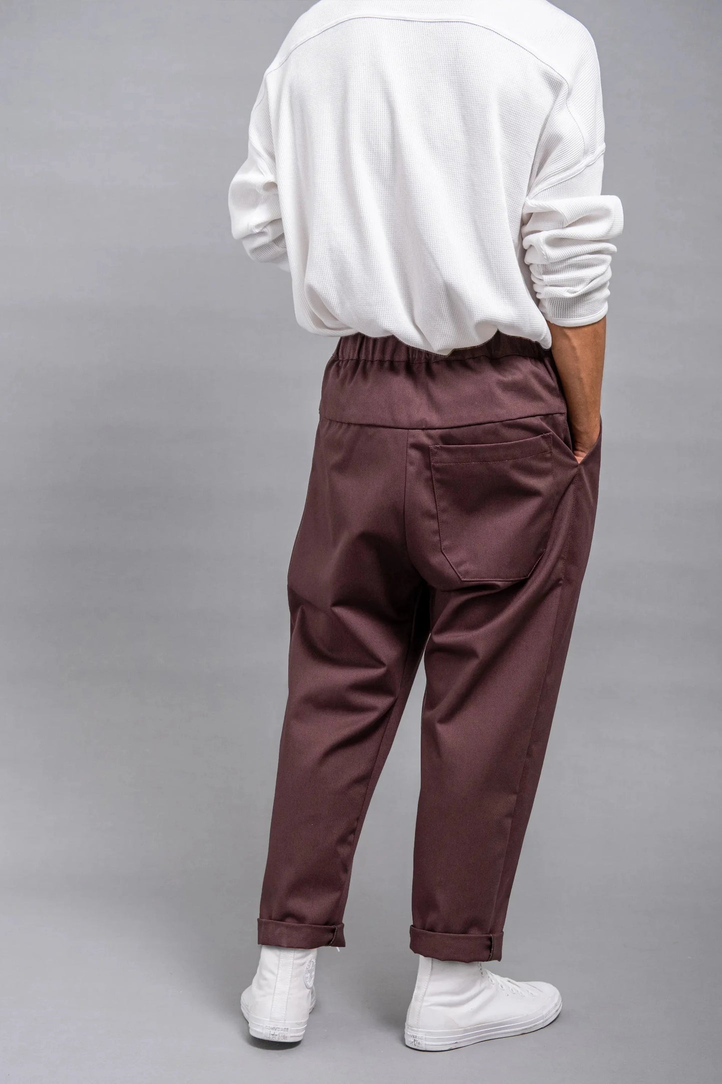 Back view of a person wearing relaxed fit burgundy pleated pants with an elastic waistband and cuffed hems, paired with a loose white long-sleeve shirt and white shoes.