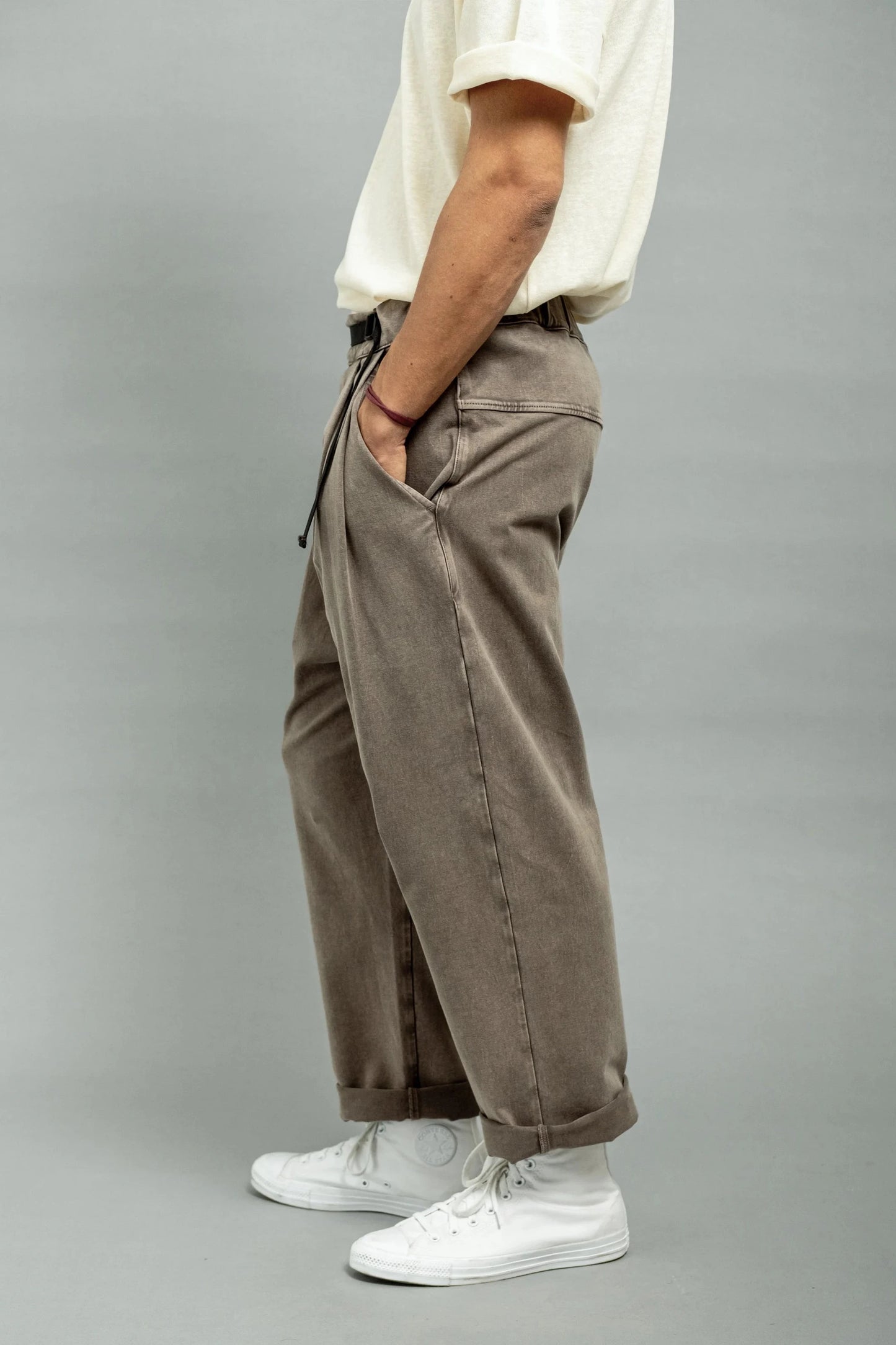 Side view of a person wearing relaxed fit brown double-knit pants paired with white high-top sneakers and a light-colored short sleeve shirt against a plain background.
