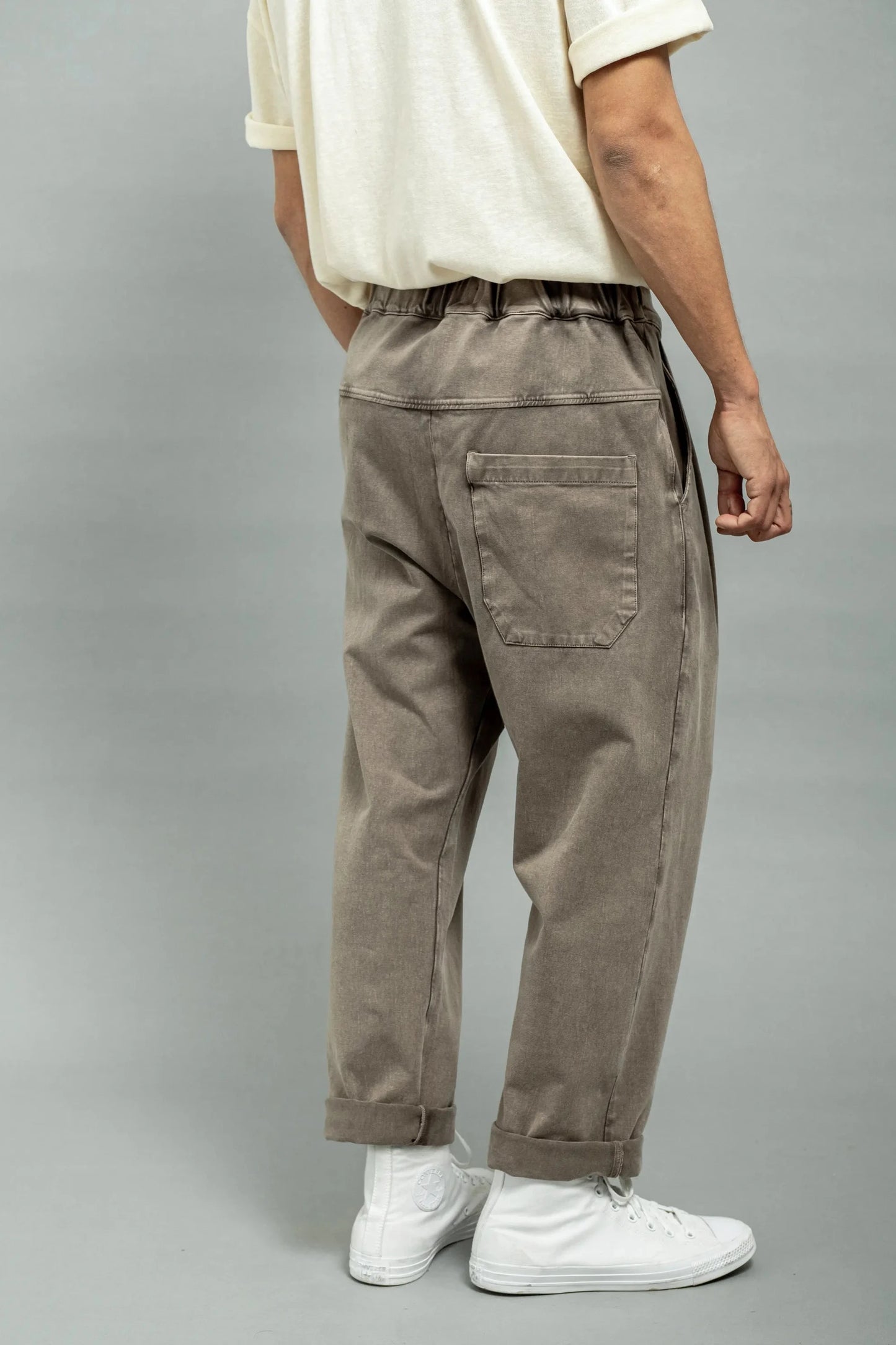 Back view of relaxed fit men's pants in washed coffee brown fabric with elastic waistband and cuffed hems, styled with white sneakers and a cream short-sleeve shirt.