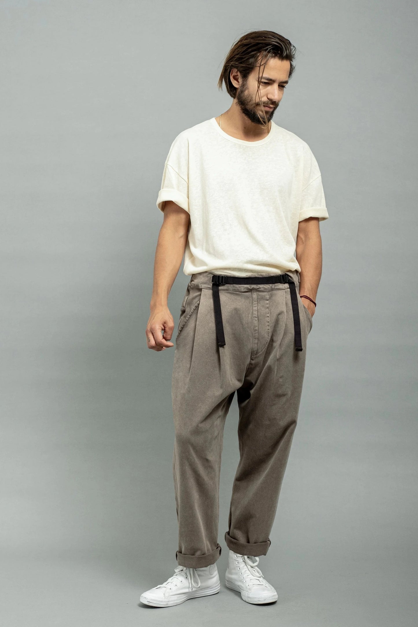 Man wearing relaxed-fit pleated pants with a black belt, paired with a loose white short-sleeve shirt and white high-top sneakers, standing against a gray background.