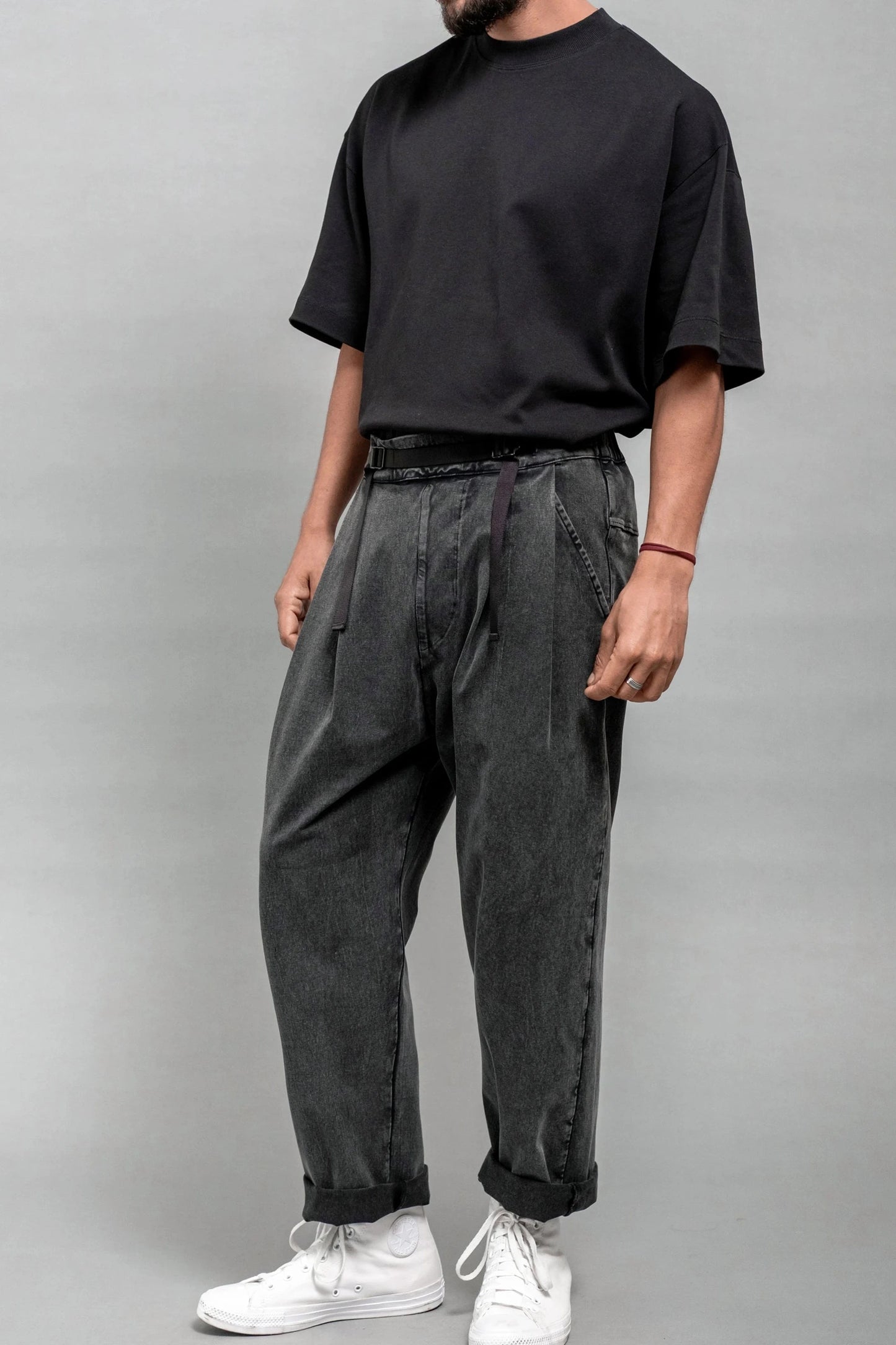 Man wearing relaxed fit pleated black wash double-knit pants paired with a loose black t-shirt and white sneakers against a plain gray background