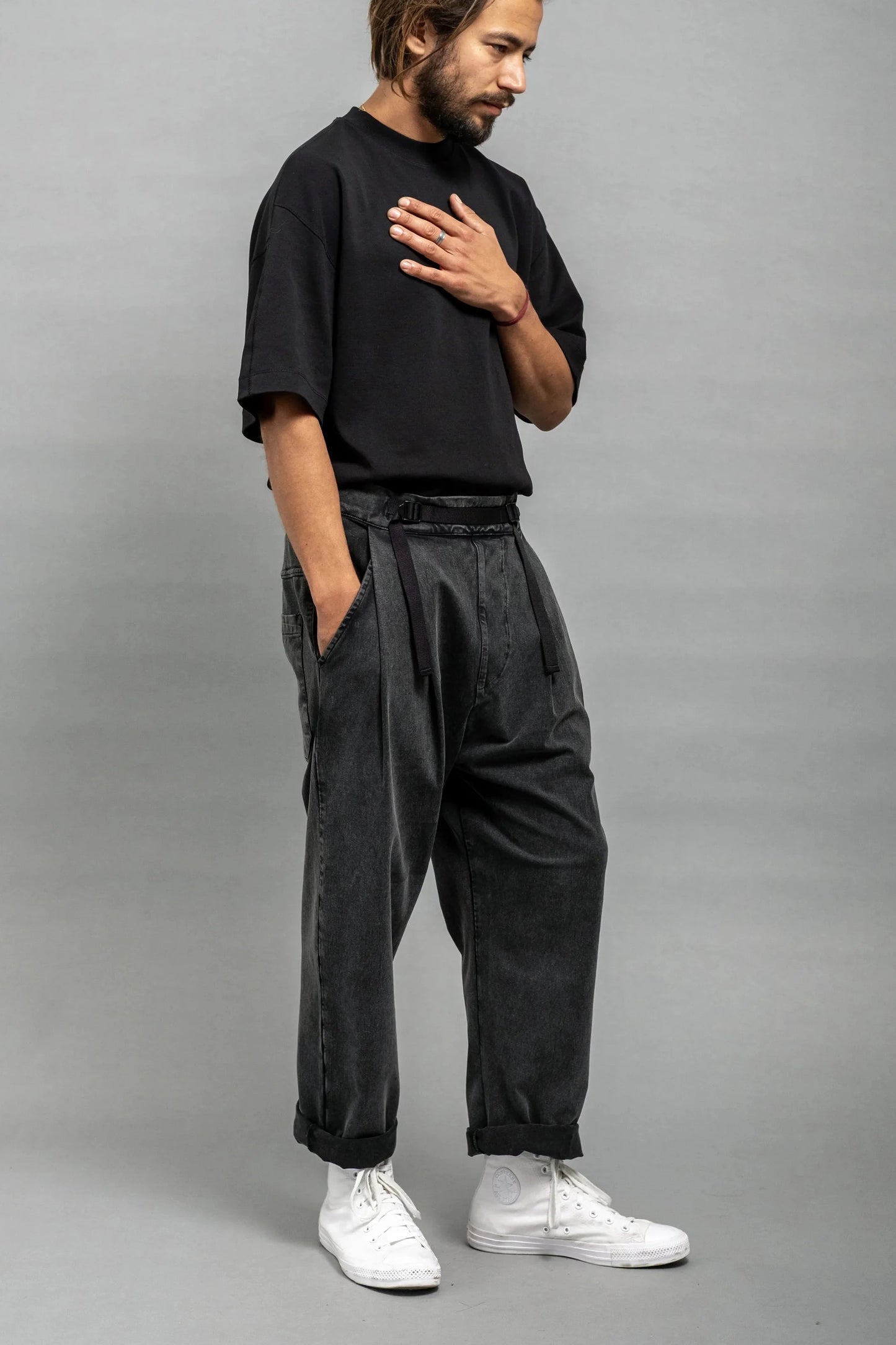 Man wearing relaxed fit pleated black wash double-knit pants with a casual black t-shirt and white high-top sneakers against a plain gray background.
