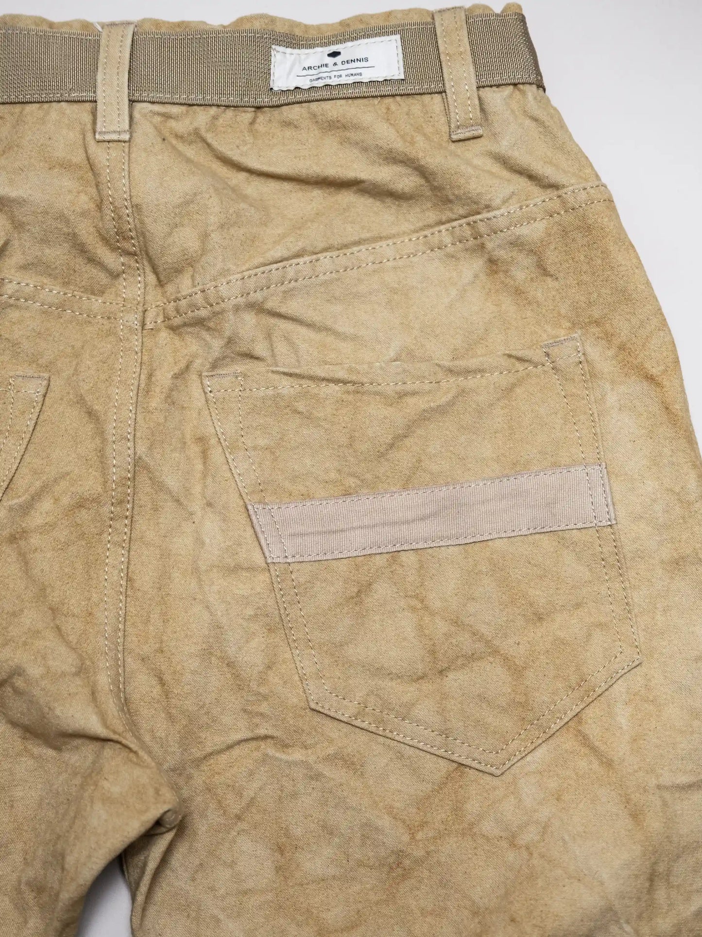 Close-up of beige utility-style pants made from textured deadstock fabric, featuring rear pockets with reinforced detailing and an olive green woven belt looped through the waistband.