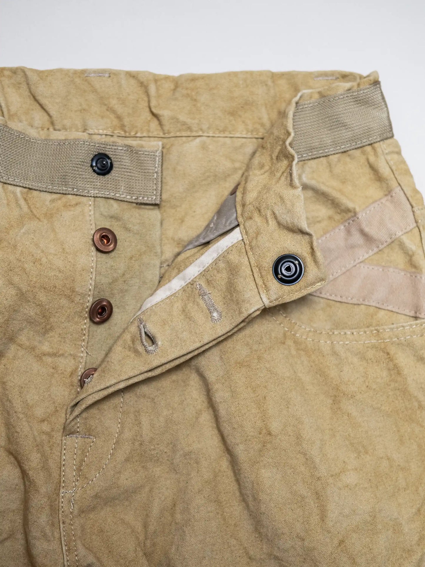 Close-up of beige utility pants featuring a button fly closure, reinforced waistband, and detailed stitching on soft fabric with a worn, vintage-inspired texture.