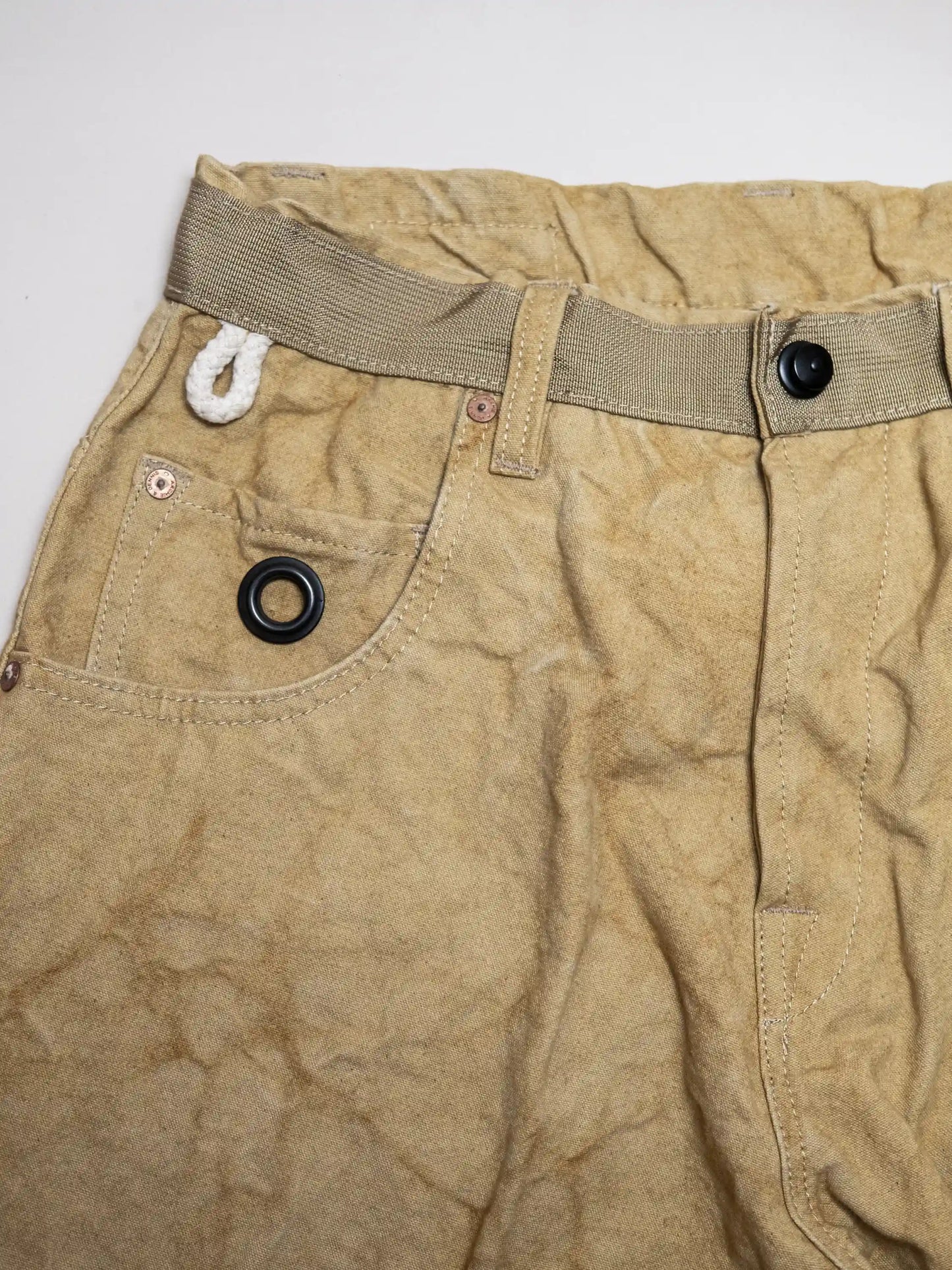 Close-up view of beige utility style pants made from textured vintage fabric, featuring a built-in fabric belt, cargo pocket with black grommet, and contrast white rope loop detail.