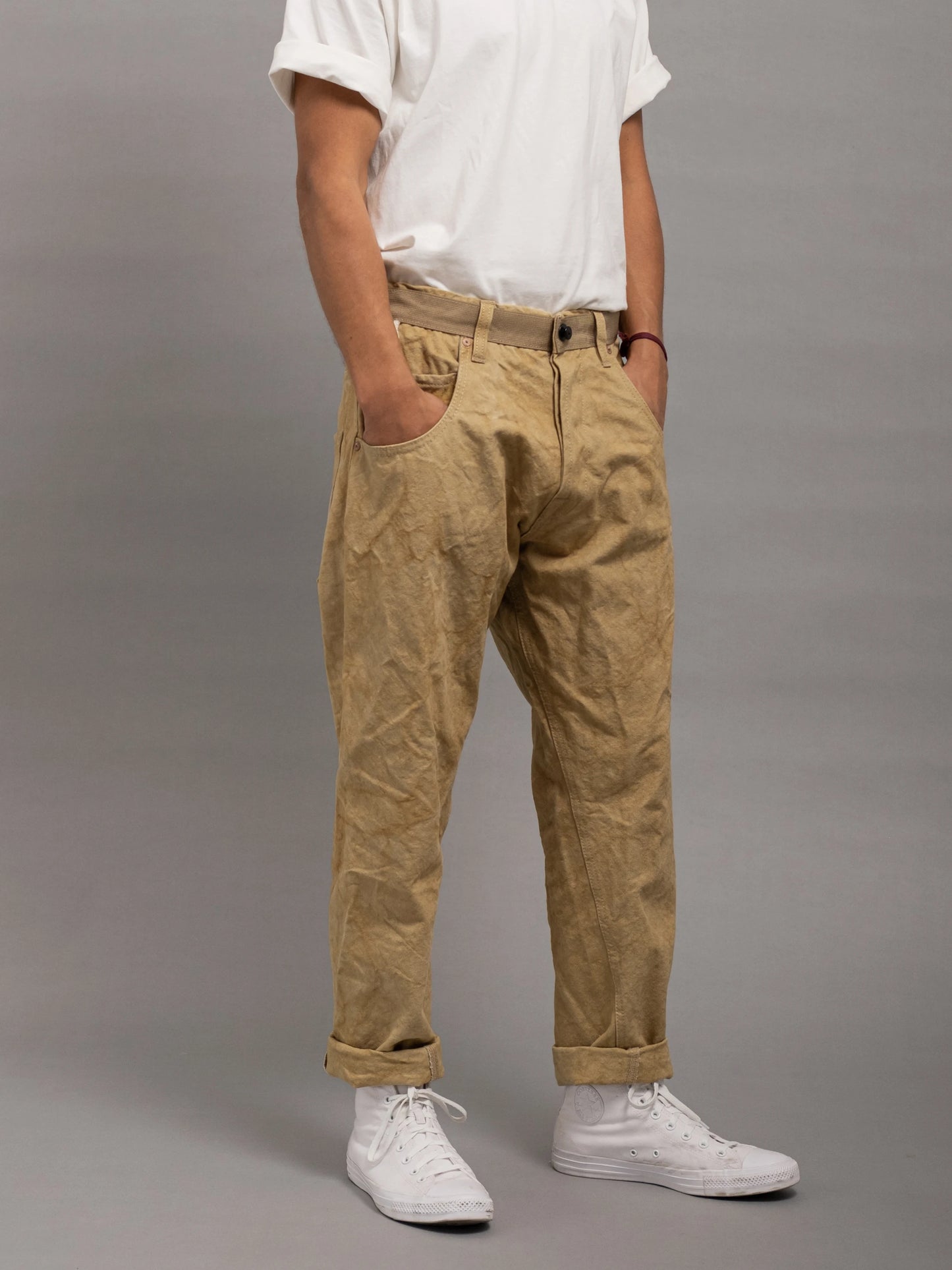 Man wearing relaxed fit pleated tan utility-style pants paired with a white rolled-up sleeve t-shirt and white sneakers against a plain gray background.