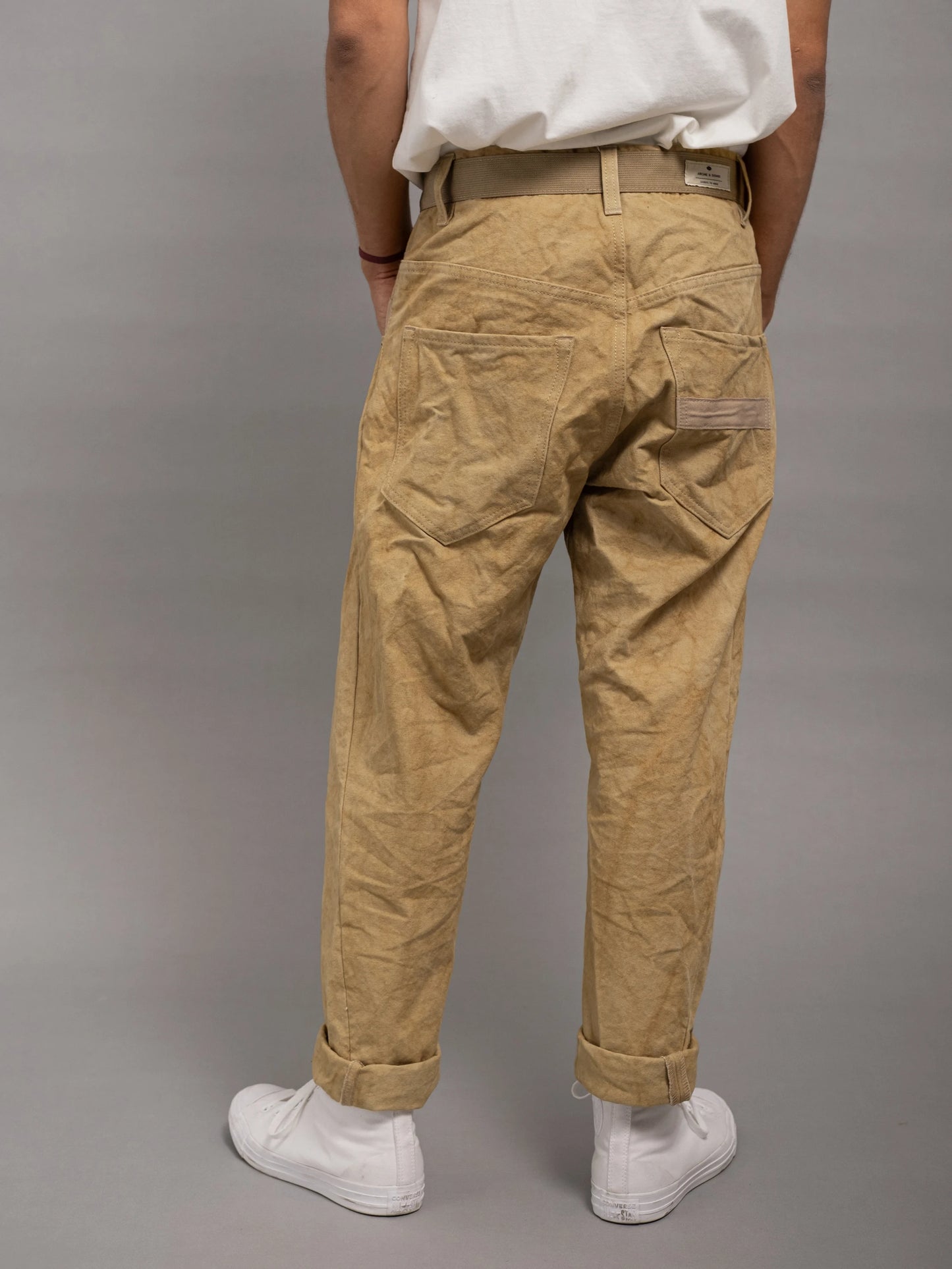 Back view of relaxed fit pleated pants in tan fabric with utility-style details, rolled cuffs, and a beige belt, worn with a white t-shirt and white sneakers.