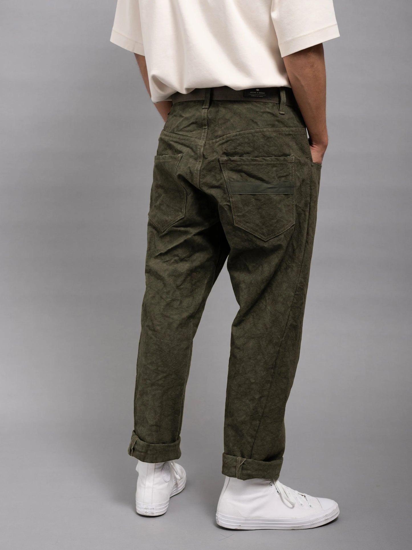 Rear view of relaxed fit olive green utility style pants with pleated details, worn with a casual white shirt and white high-top sneakers.