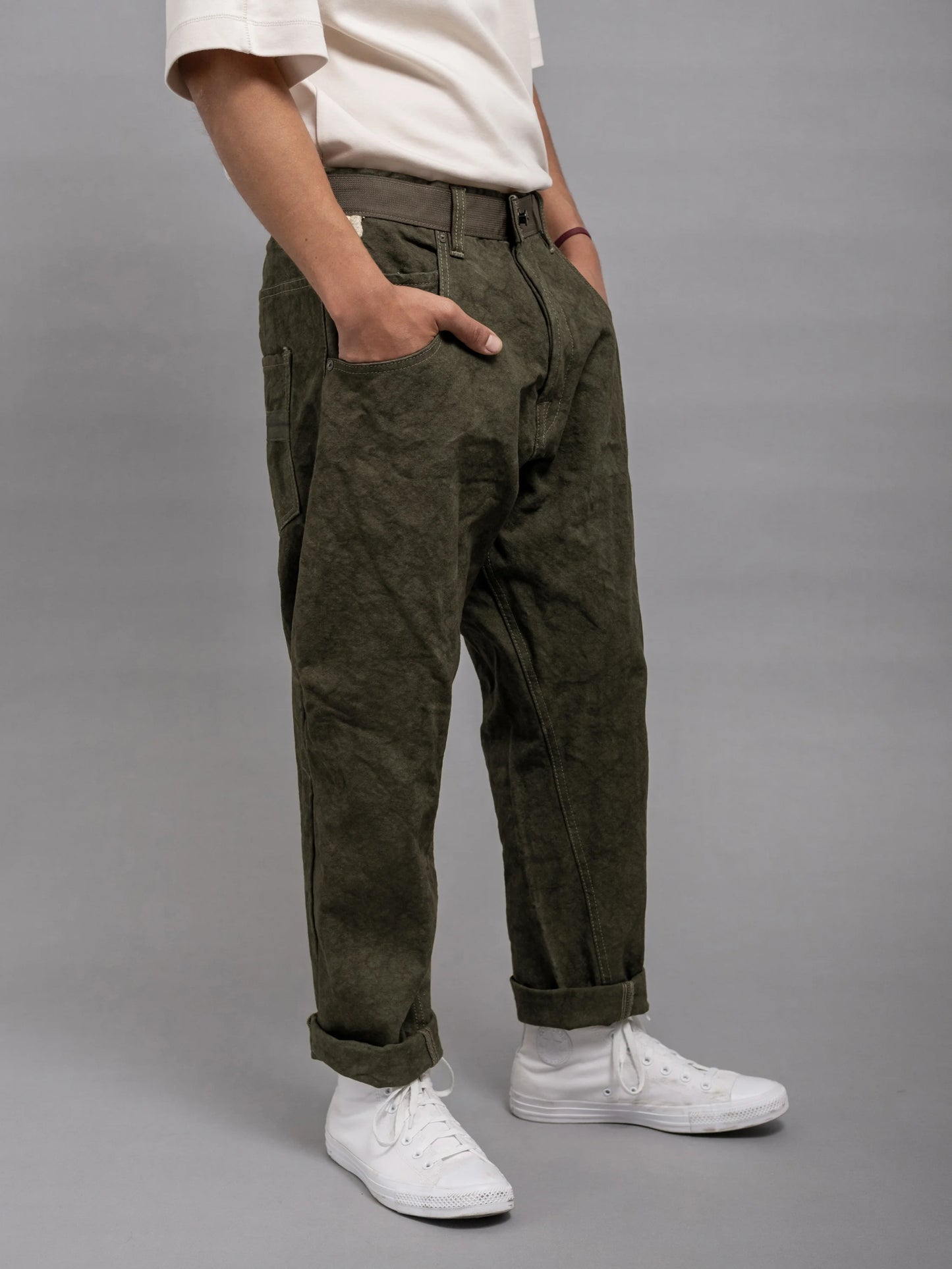 Side view of a person wearing olive green relaxed fit pleated utility style pants paired with white sneakers and a white t-shirt, standing against a plain gray background.