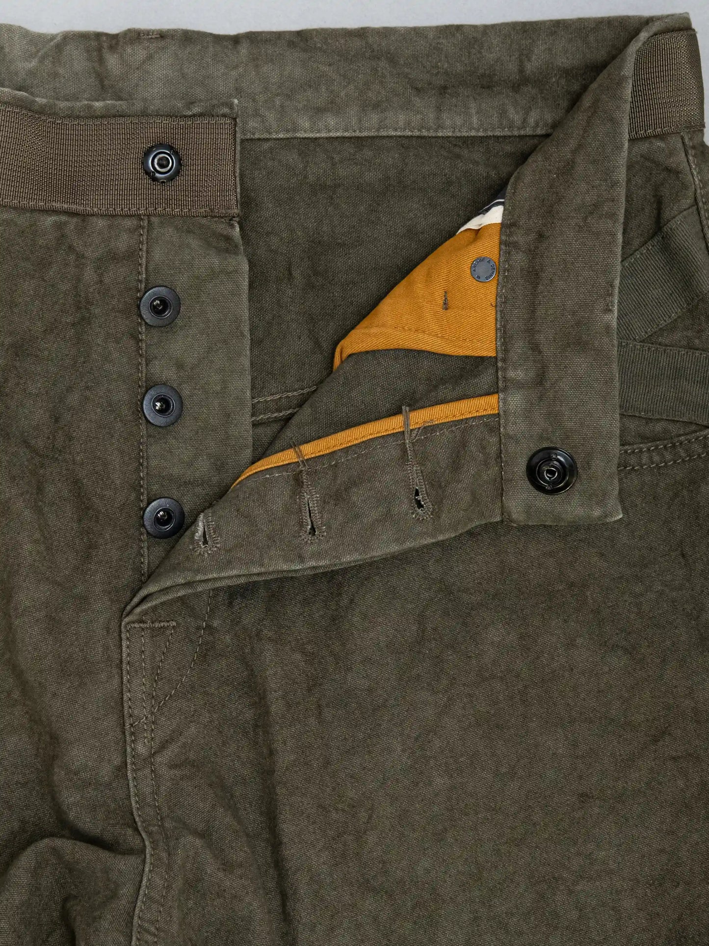 Close-up of olive green relaxed fit pants with button fly and contrasting mustard yellow inner fabric detail, showcasing sturdy stitching and utility-inspired design.