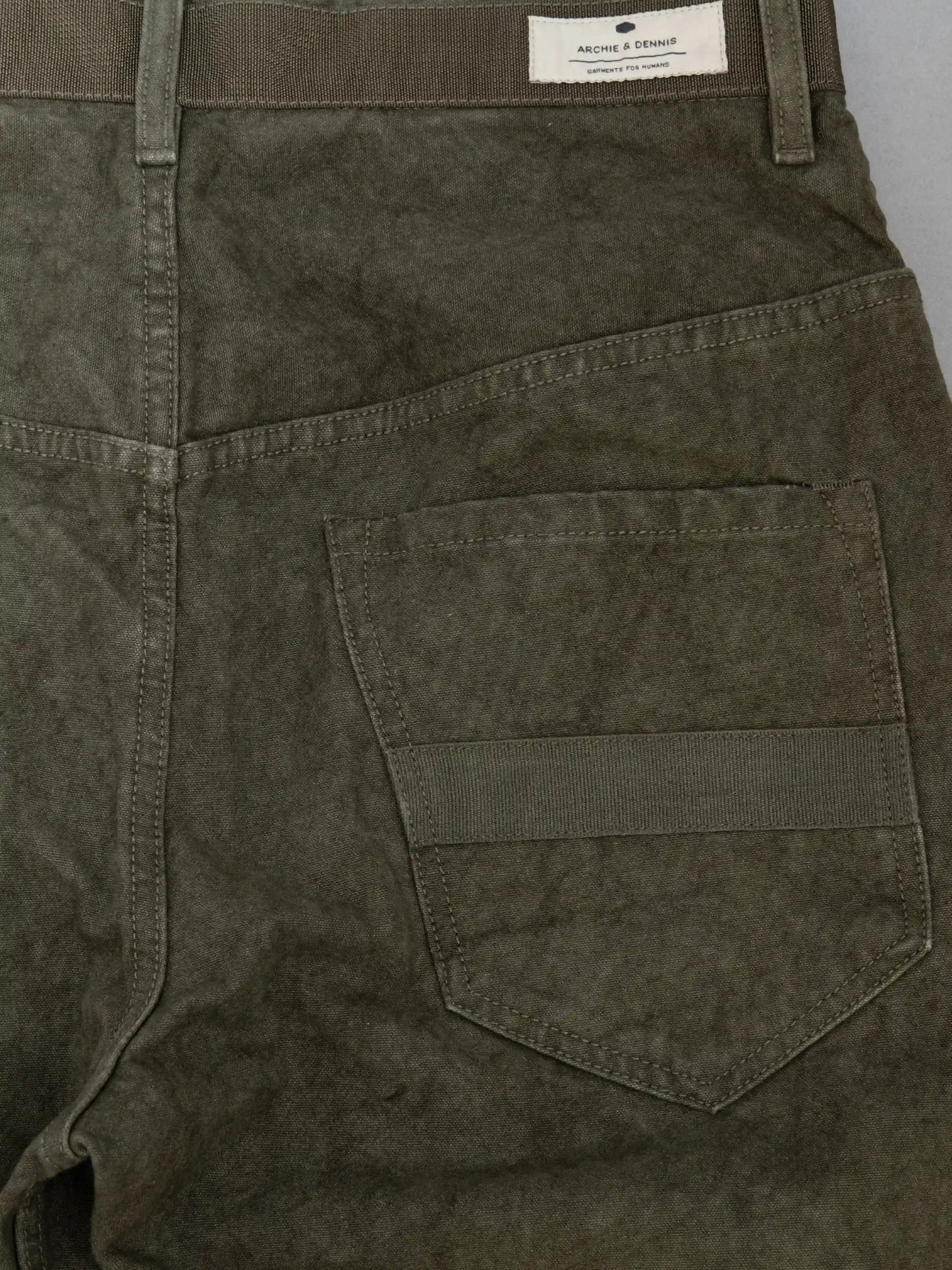 Close-up of olive-colored utility style pants made from textured fabric featuring a back pocket with a horizontal strap detail and a branded label on the waistband.