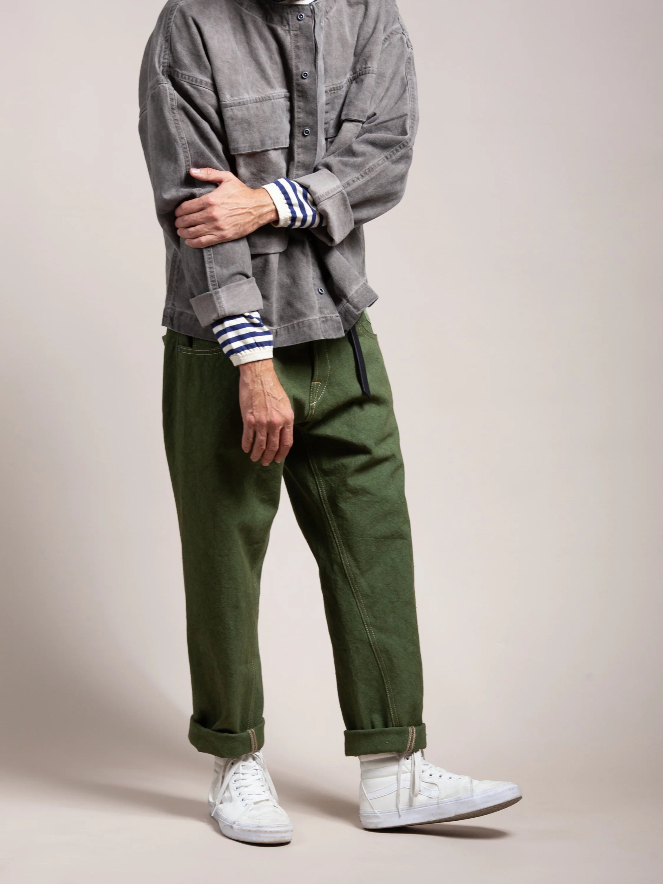 Man wearing olive green relaxed fit pants rolled at the cuffs paired with a gray jacket over a striped long sleeve shirt and white sneakers against a plain neutral background.