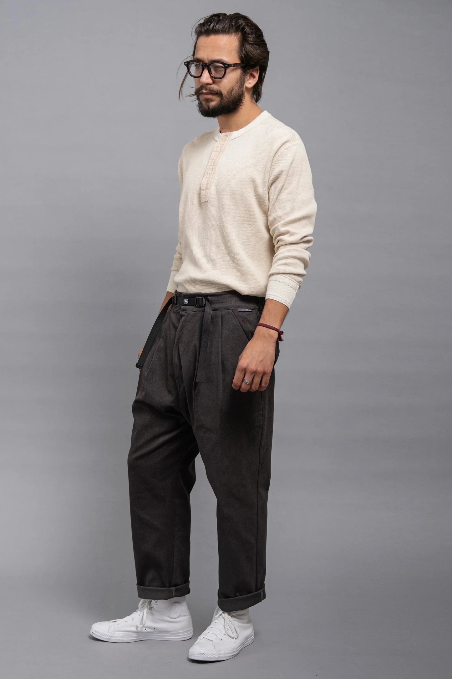 Man wearing relaxed fit pleated grey twill pants paired with a cream long-sleeve henley shirt and white sneakers against a plain grey backdrop.