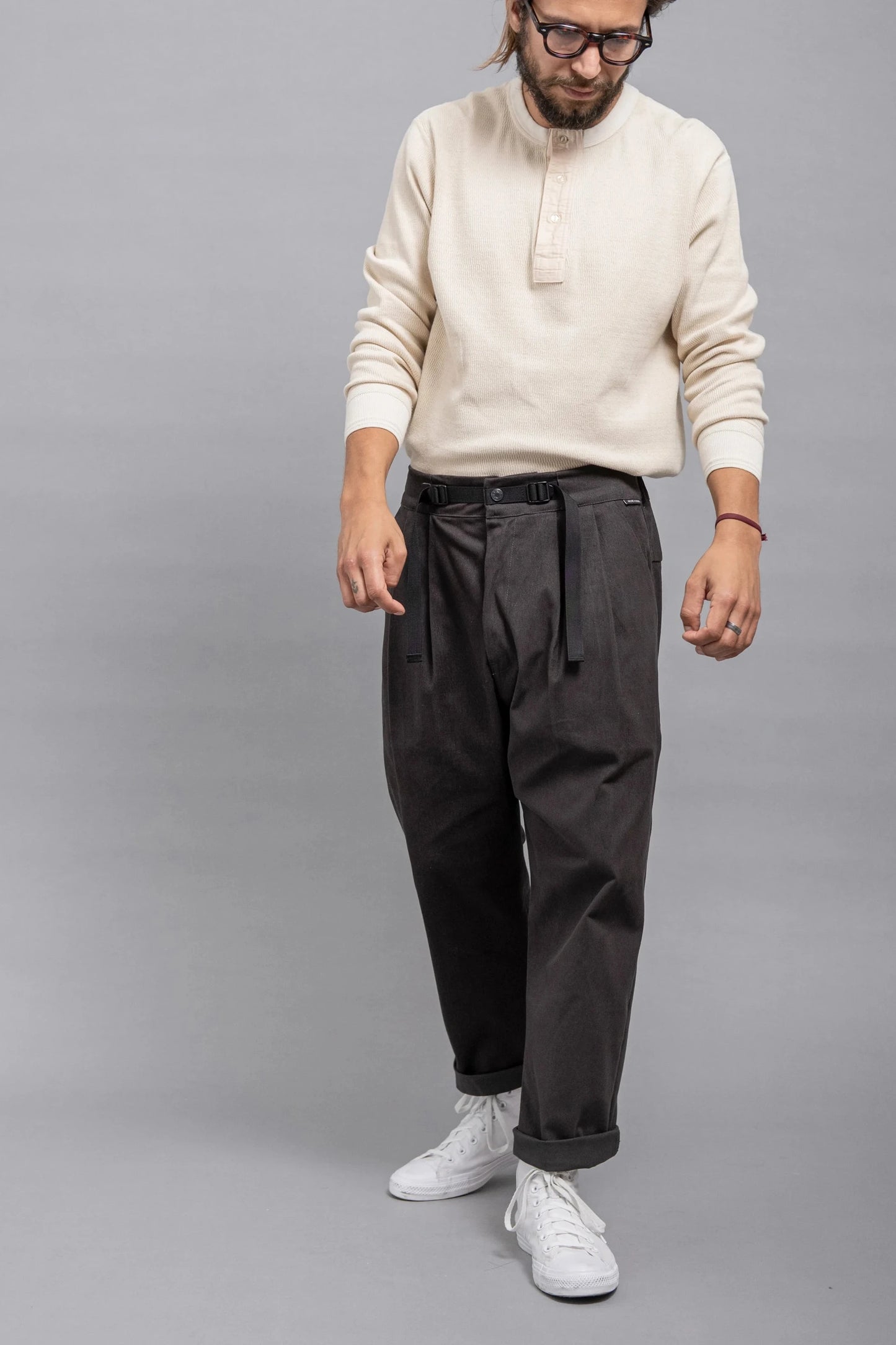 Man wearing relaxed fit pleated dark grey twill pants paired with a cream long-sleeve henley shirt and white sneakers against a plain grey background.