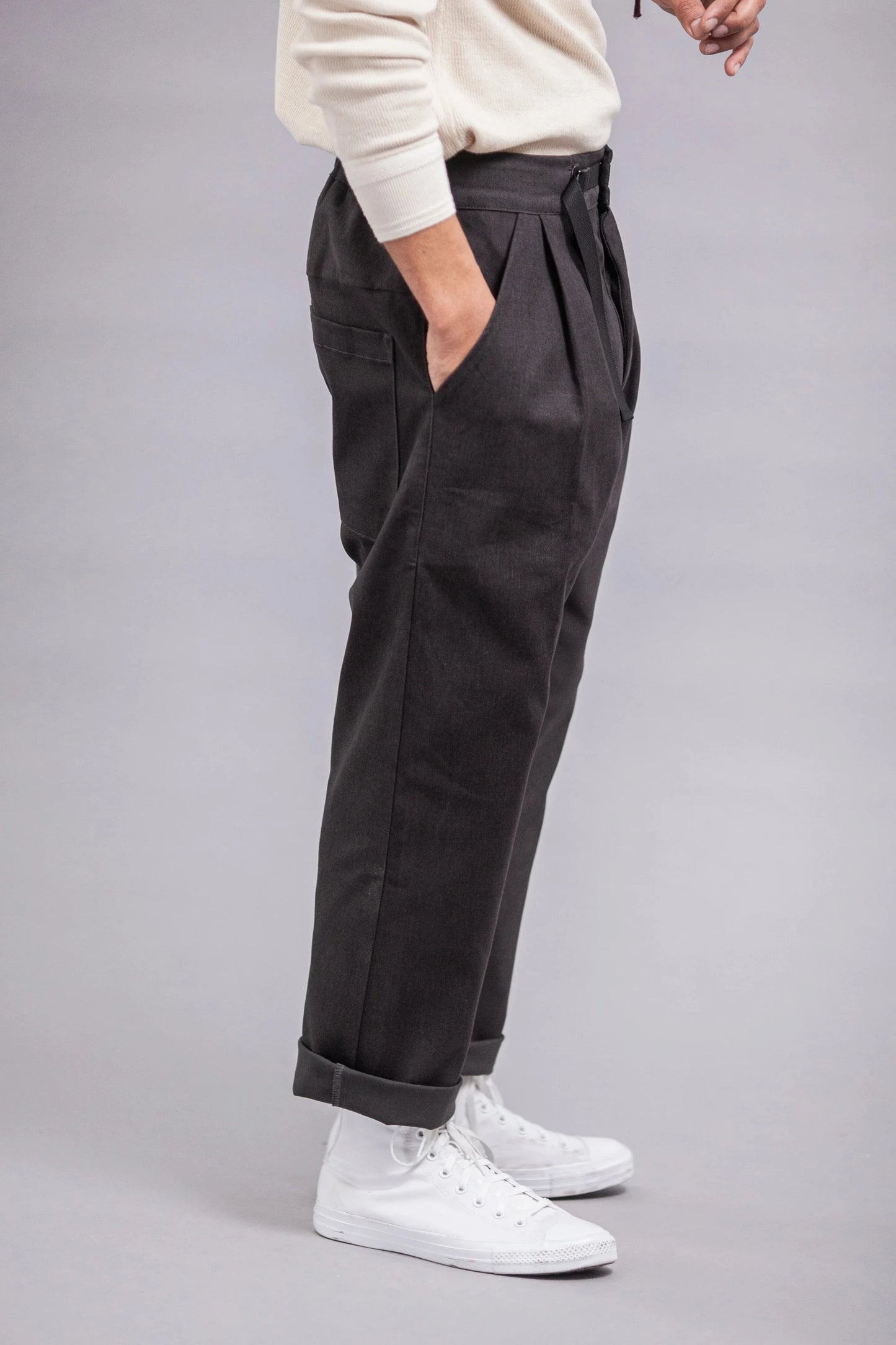 Side view of a person wearing relaxed fit pleated grey twill pants with rolled cuffs paired with white high-top sneakers and a light-colored long sleeve top.