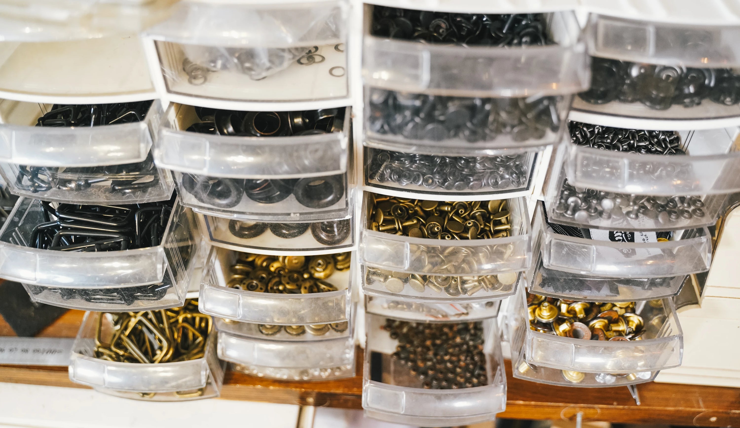 Clear plastic drawers filled with various small metal hardware pieces including rings, snaps, and eyelets in black, gold, and silver colors, organized for easy access.
