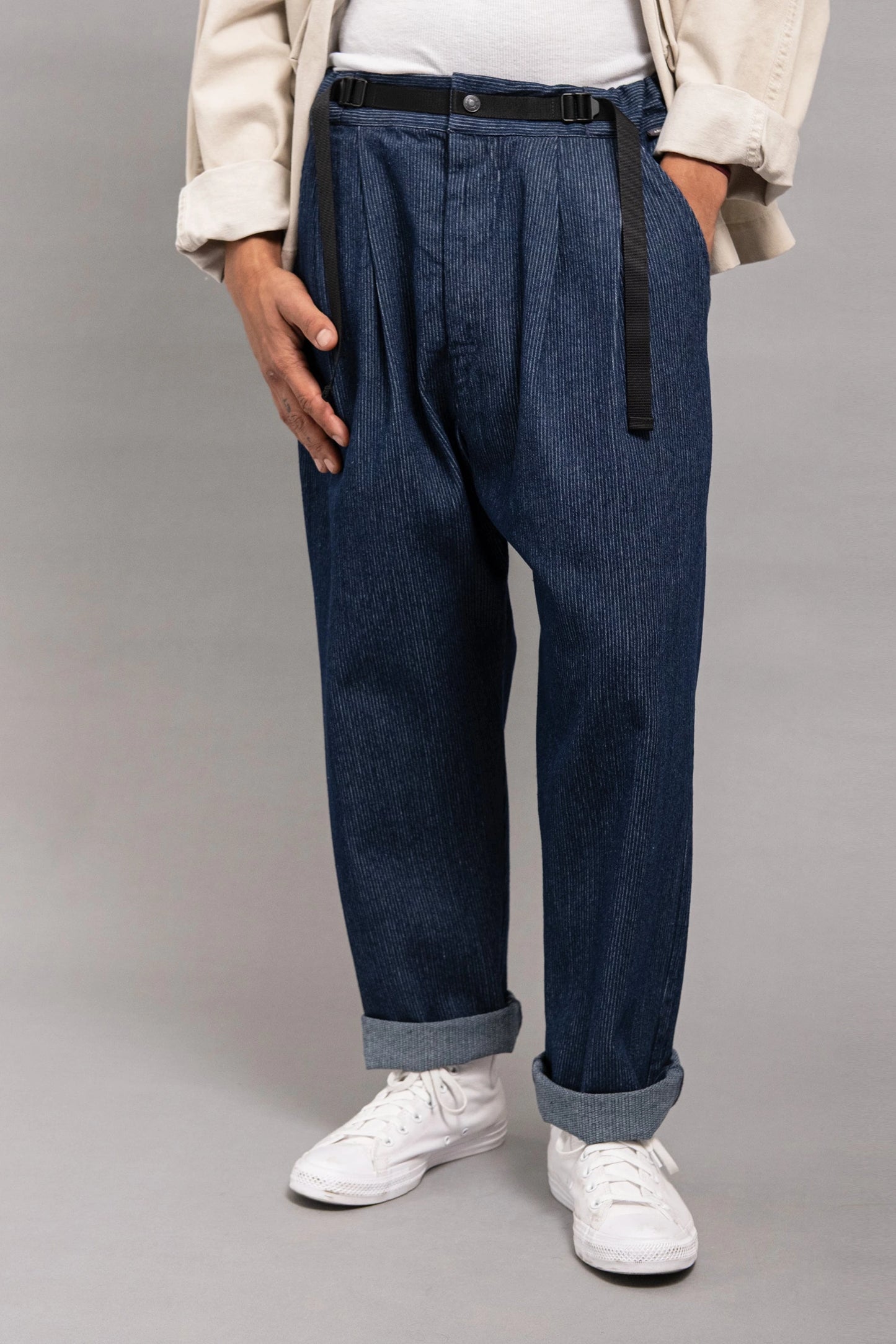 Front view of a person wearing relaxed fit pleated indigo pants with a black belt, cuffed hems, white sneakers, a white shirt, and a light beige jacket.