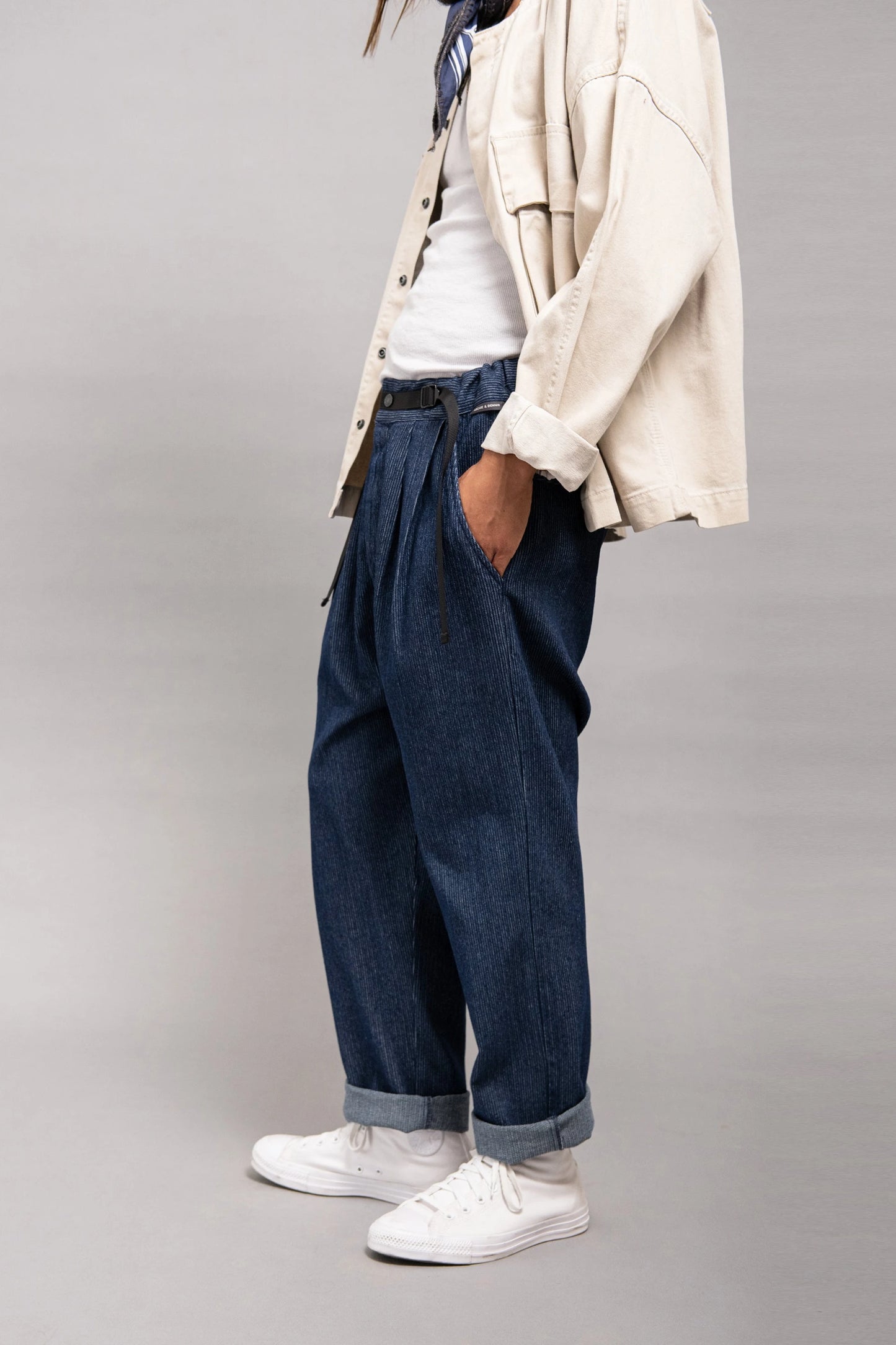 Side view of a person wearing relaxed fit indigo denim pants with pleats and rolled cuffs, paired with a casual beige overshirt, white sneakers, and a white inner shirt.