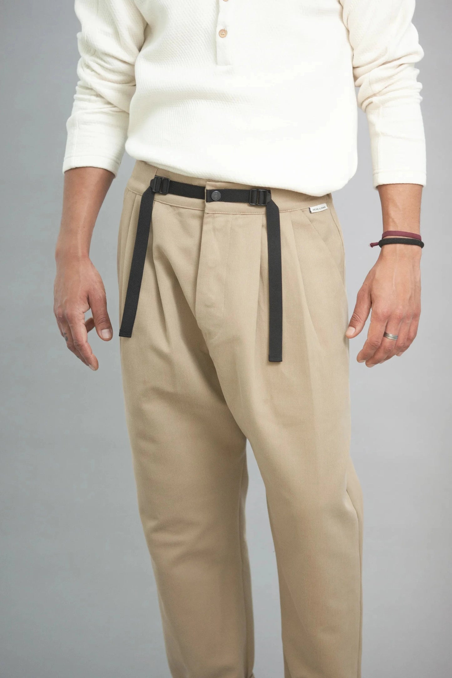 Man wearing relaxed fit pleated beige pants with a black fabric belt and an off-white long-sleeve henley shirt, styled in a minimalist, utility-inspired look.